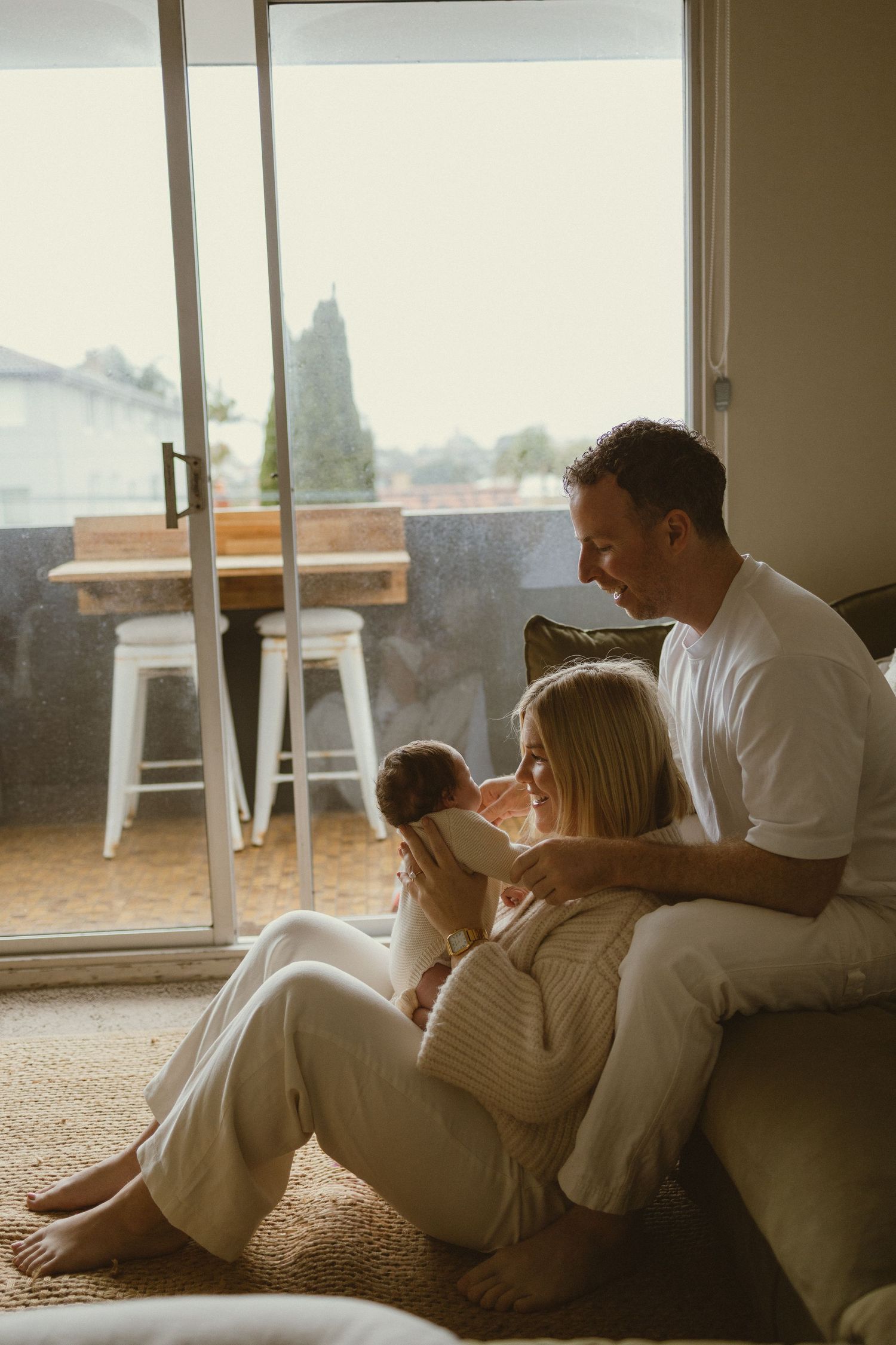 A couple sits on the floor cradling their newborn baby by a sunlit sliding glass door in a cozy living room.