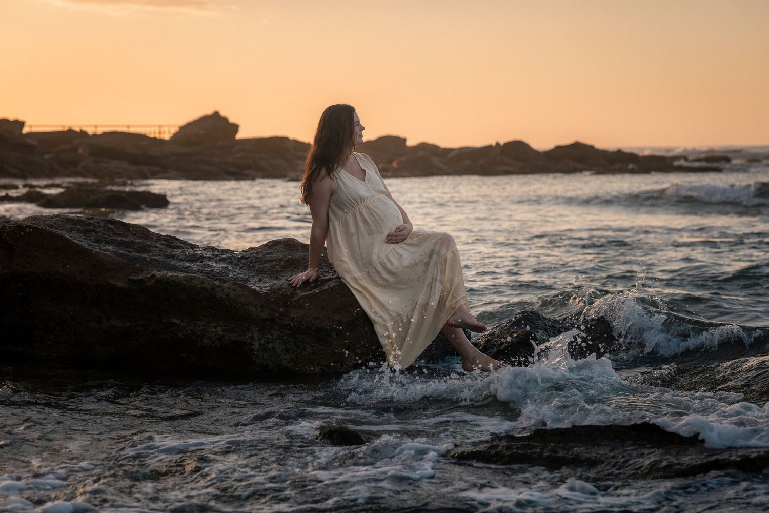 A figure in a flowing white dress stands on rocky coastal cliffs at sunset as waves crash against the shore.