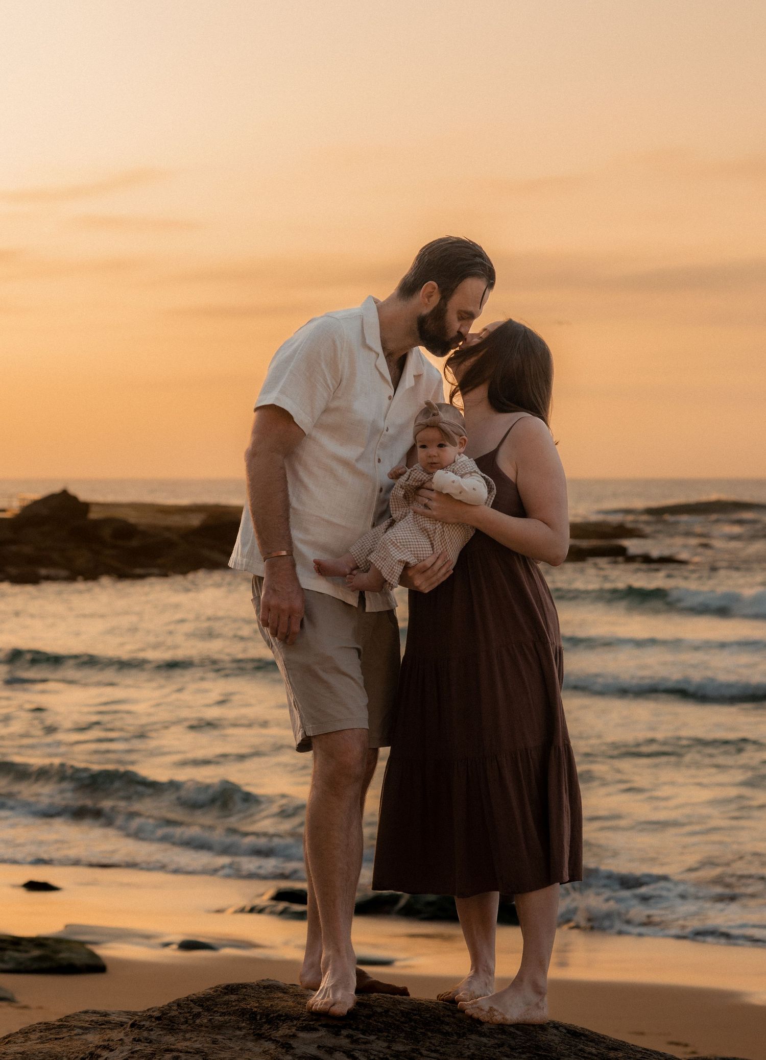 Couple shares romantic moments on a beach at sunset with ocean waves and rocky shoreline in the background.