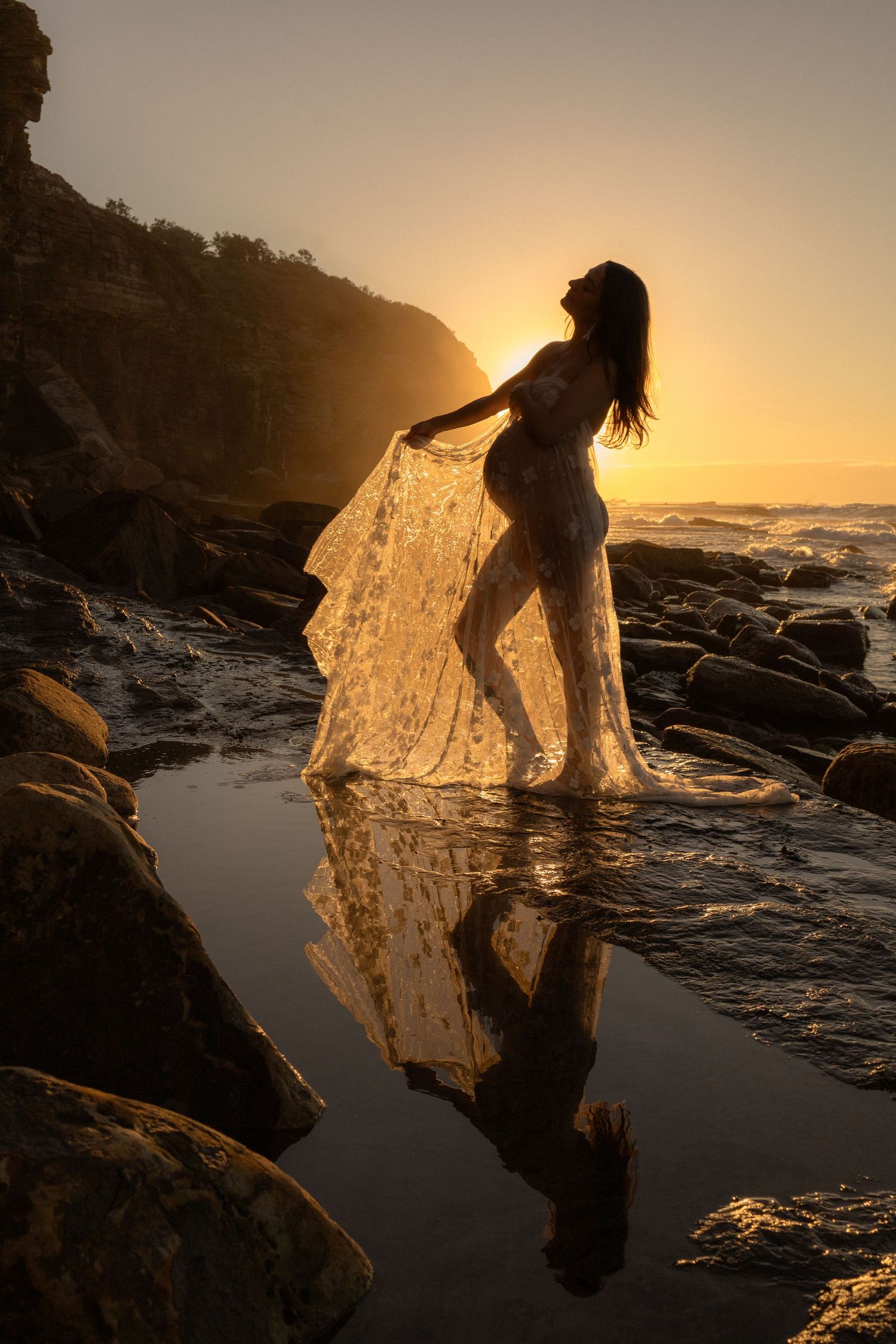 Silhouette of a woman in sheer fabric standing on rocks at golden sunset, reflected in tide pools by the ocean.
