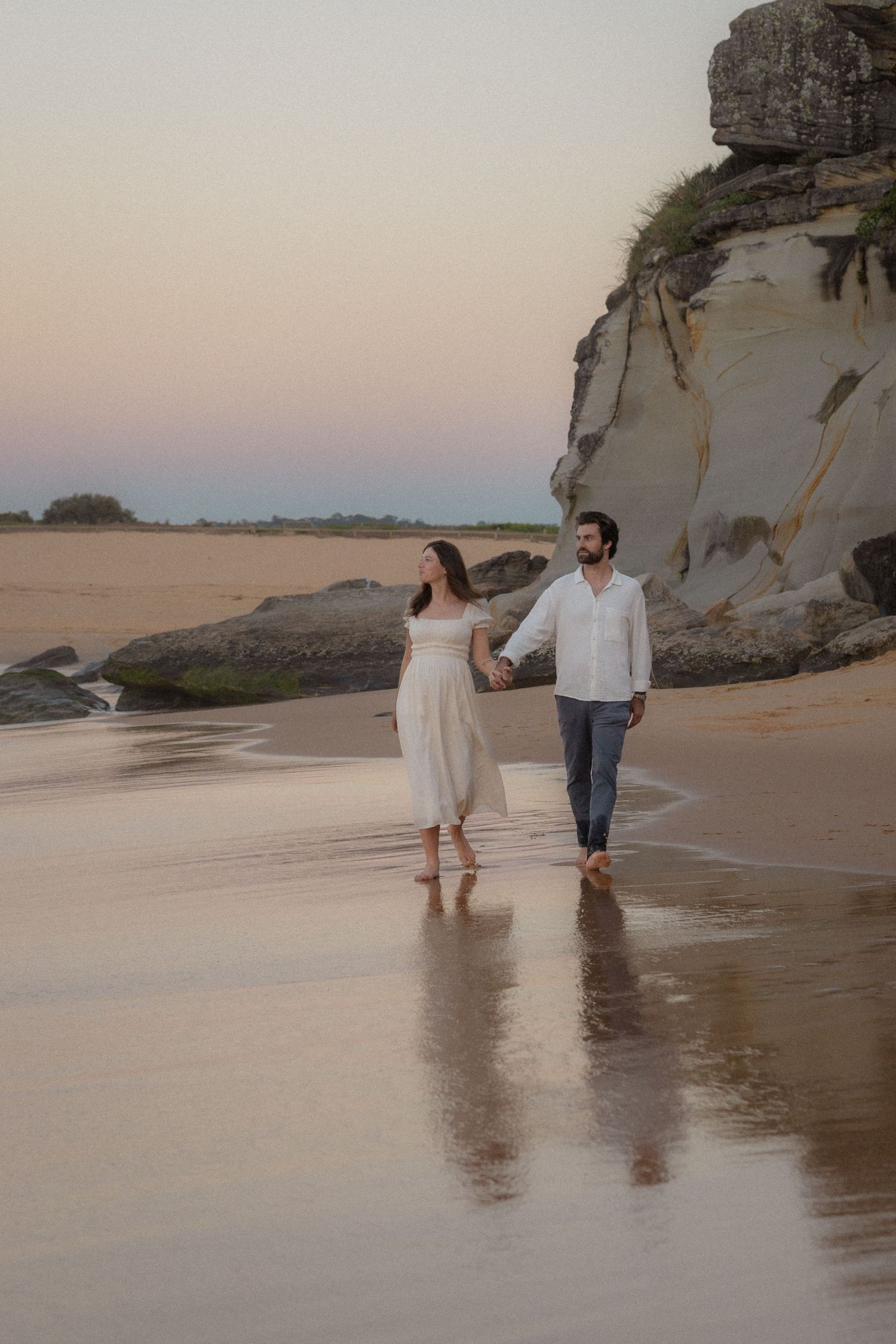 Couple holding hands walking barefoot on a reflective beach at sunset near rocky cliffs.