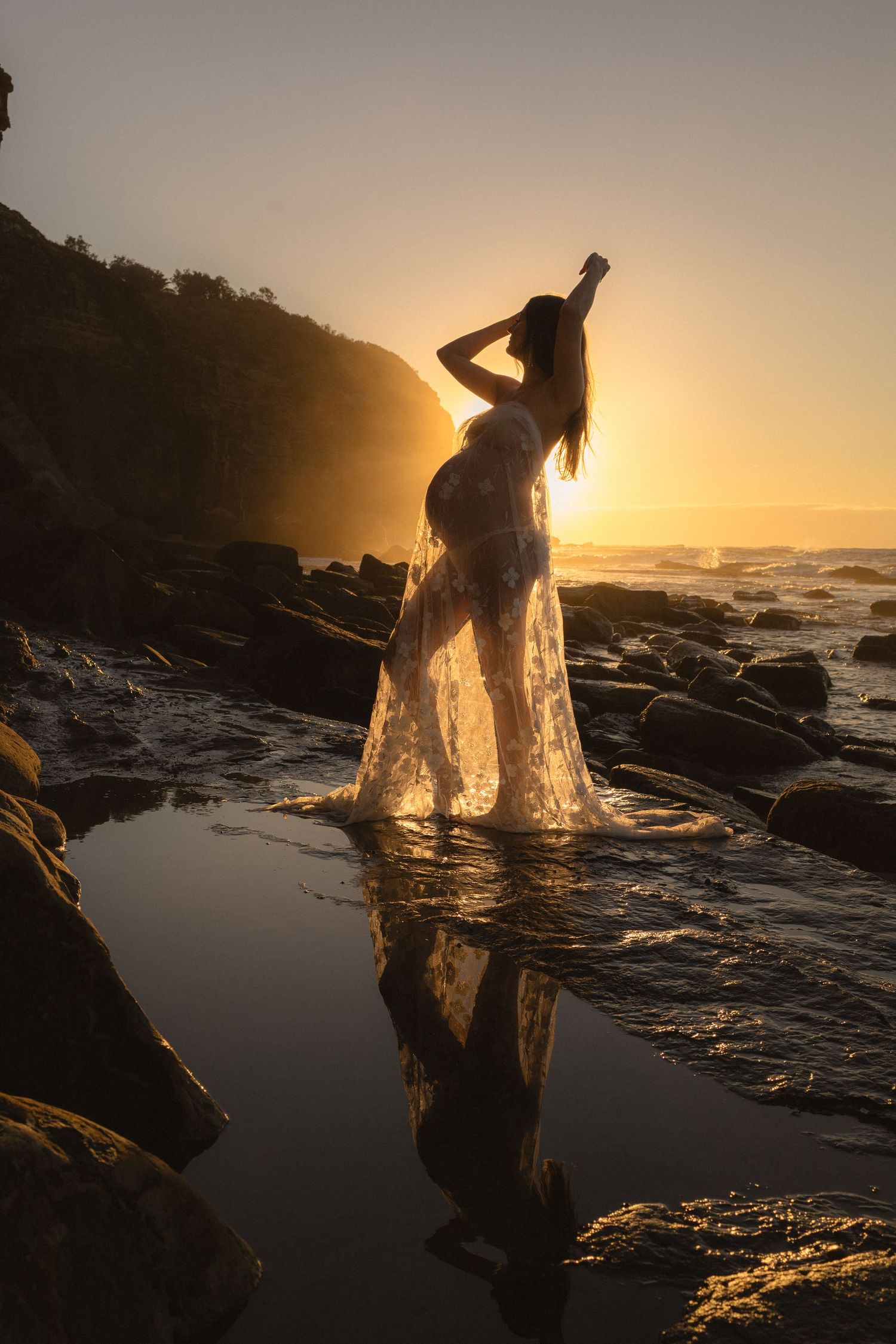 Silhouette of a pregnant woman in a flowing white dress standing on rocky beach at golden sunset with ocean reflection.