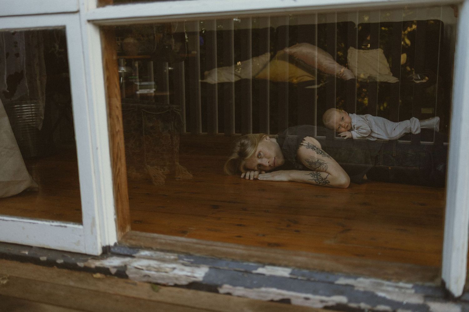Two people lying on a wooden floor, viewed through a weathered white window frame in a rustic interior.