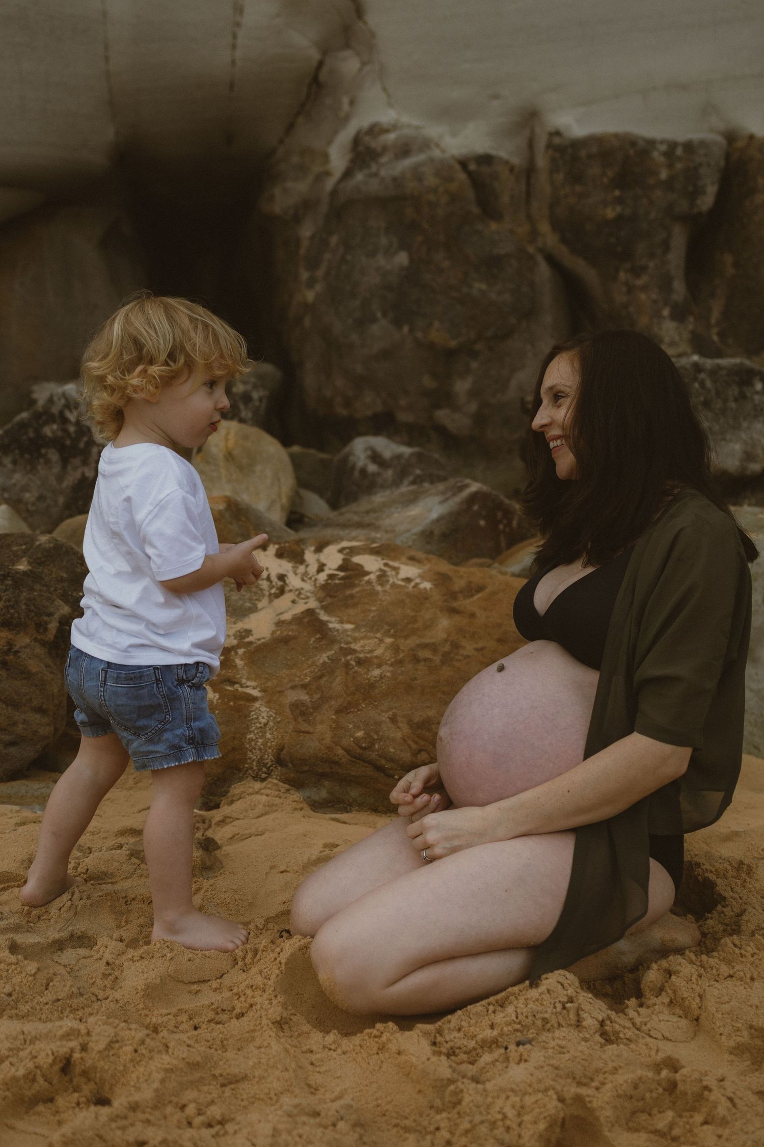 Pregnant woman in black dress sits on sandy beach smiling at curly-haired toddler in white shirt and denim shorts.
