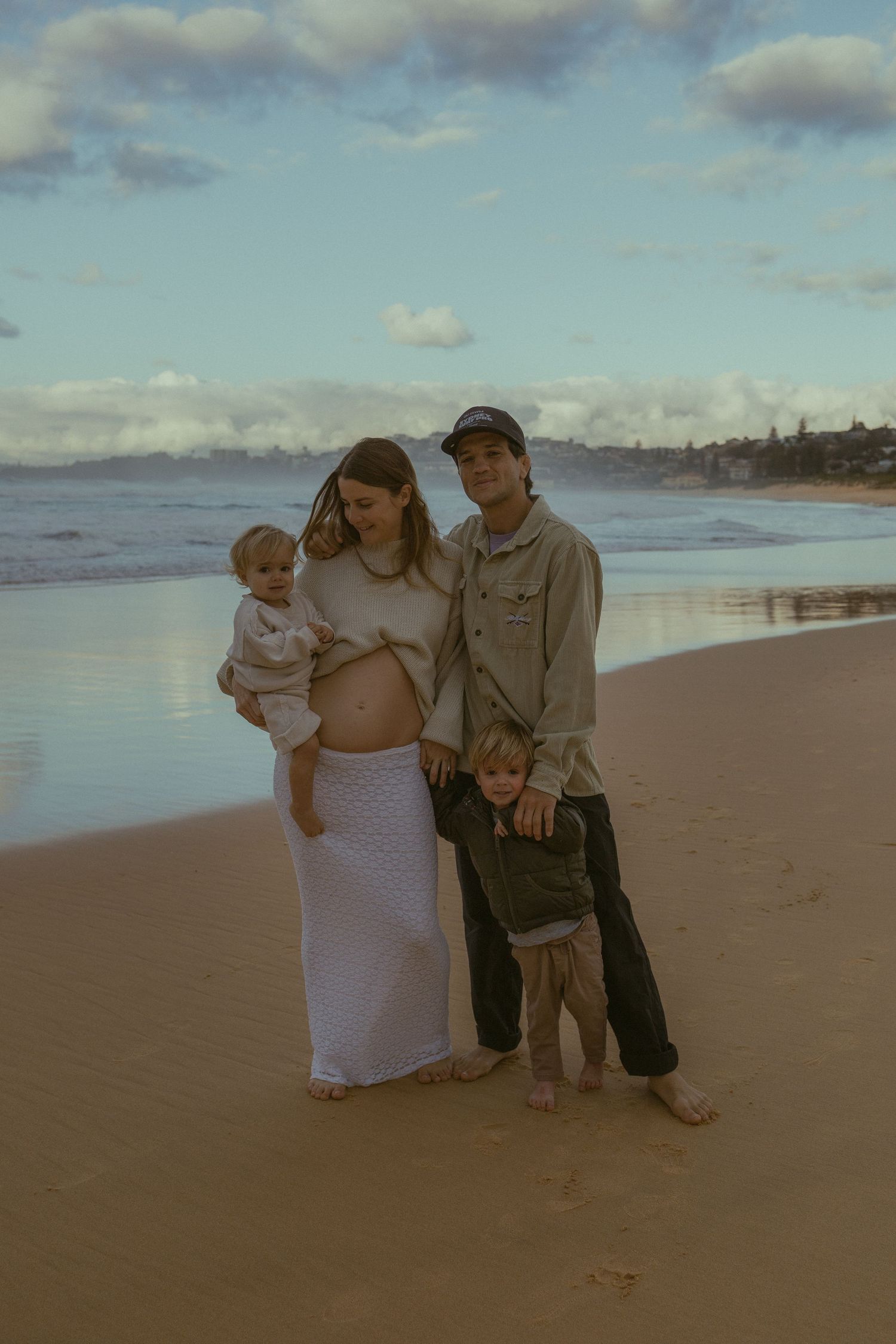 Pregnant woman holding baby with partner and toddler posing on sandy beach with ocean waves in background.