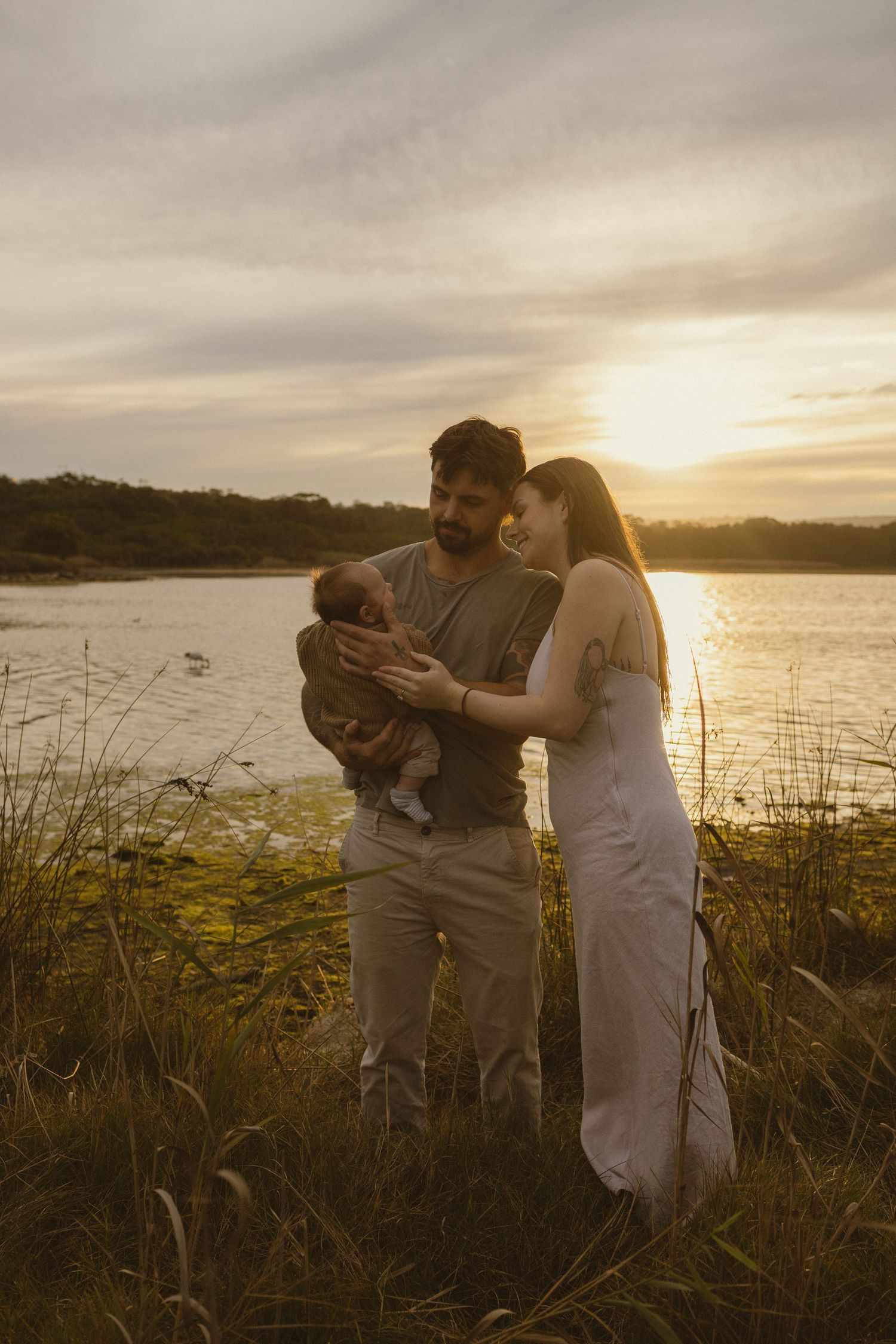 A couple admires their newborn baby outdoors by a lake at golden sunset hour.