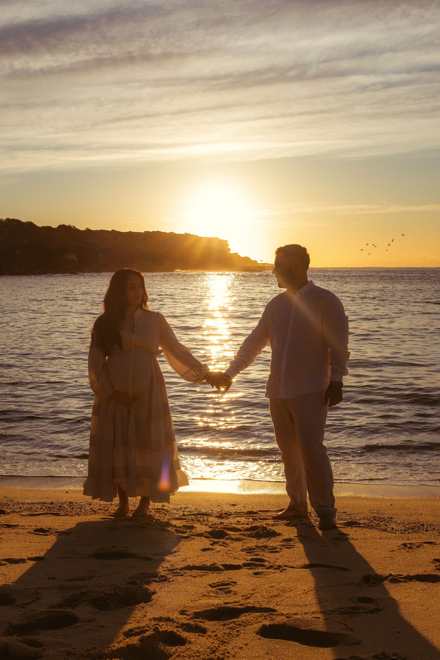 Couple holding hands on sandy beach at golden sunset, silhouetted against glowing ocean horizon.