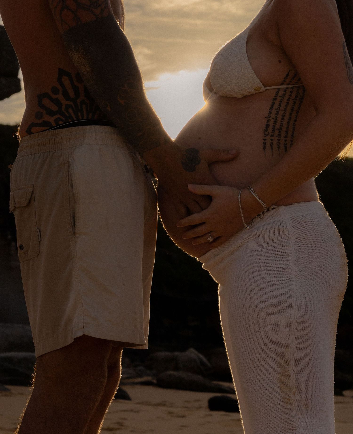 Pregnant woman in white bikini top with tattooed partner cradling baby bump on sandy beach.