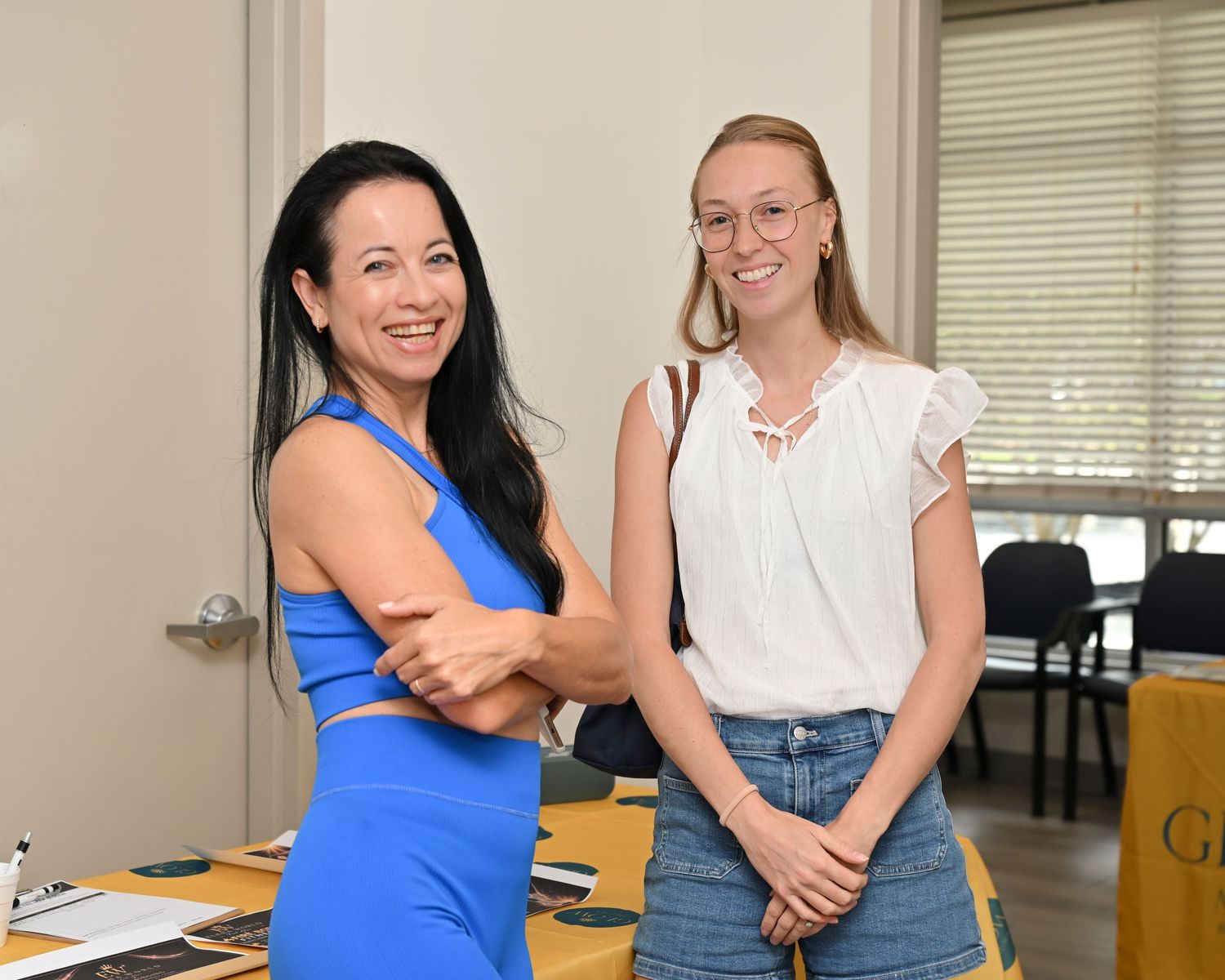 Two women smiling together in a bright room, one in a blue dress and one in a white top with jeans.