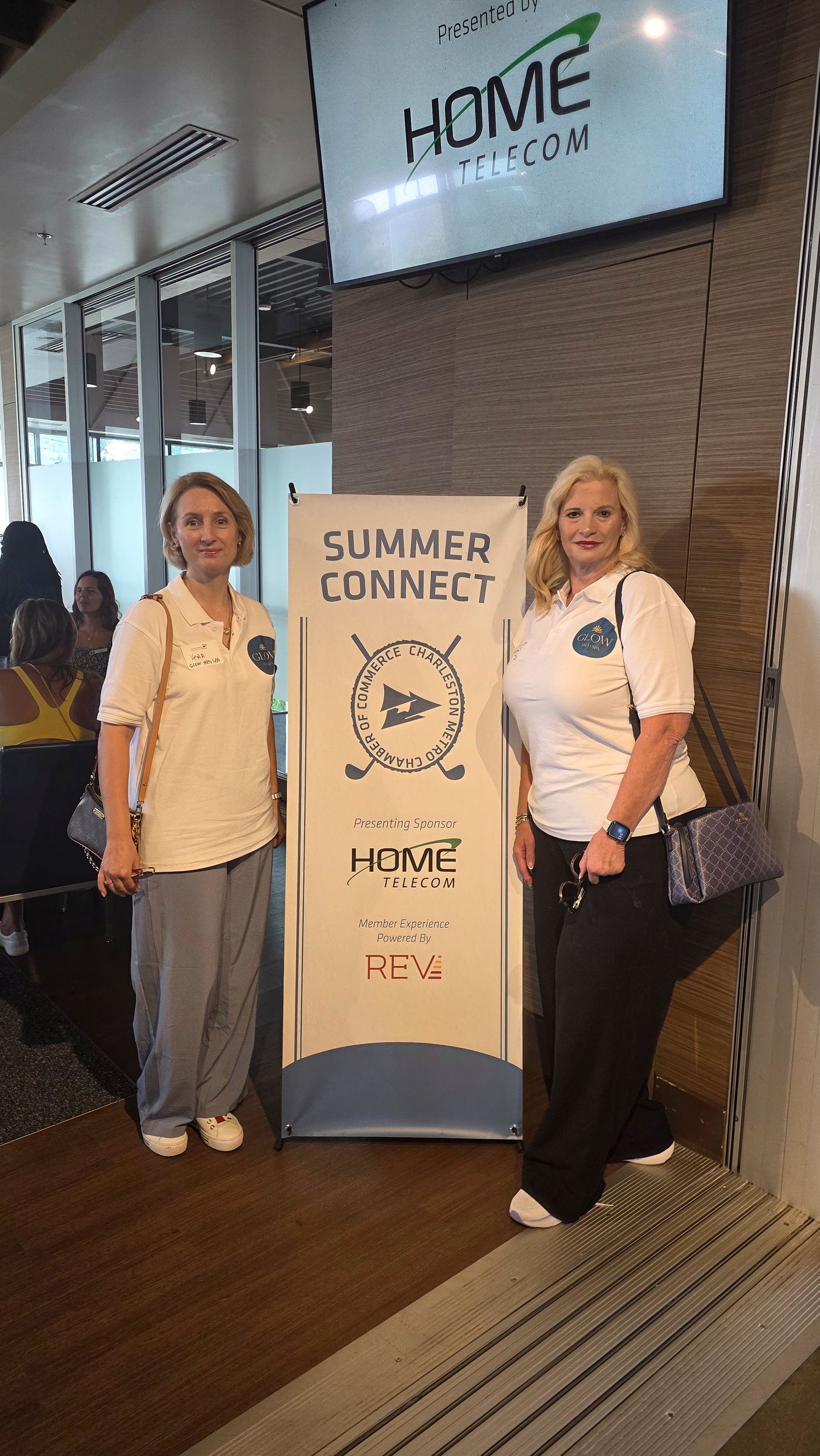 Two women in white polo shirts pose beside a Summer Connect event banner sponsored by Home Telecom at an indoor venue.