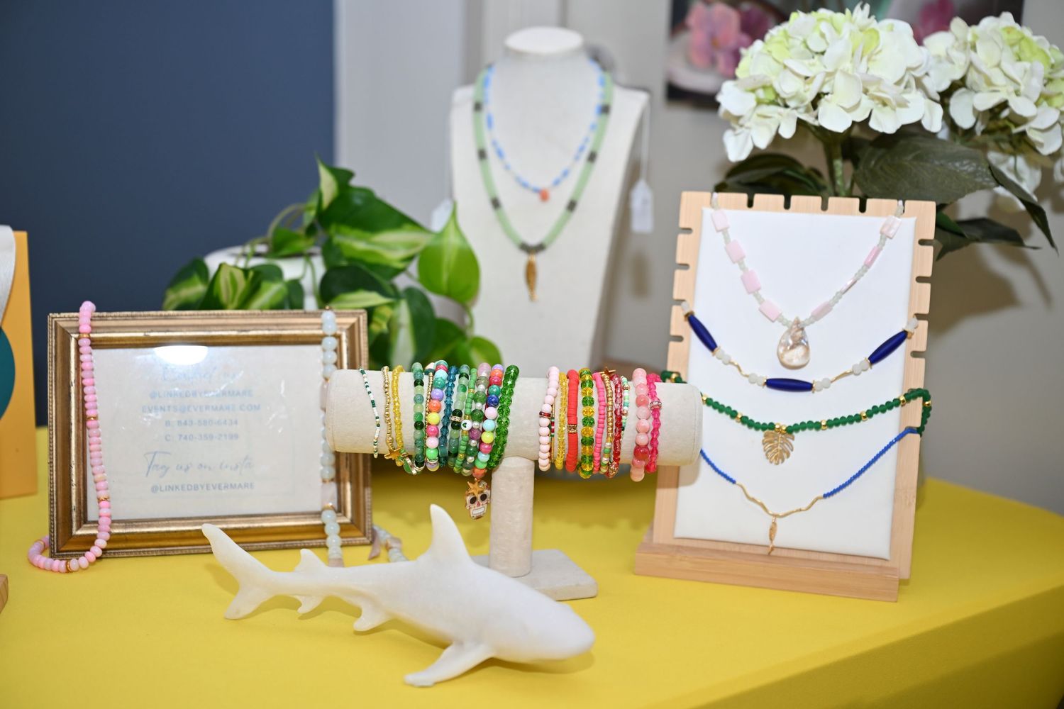 Colorful beaded necklaces and bracelets displayed on a yellow table with a white shark figurine and floral backdrop.