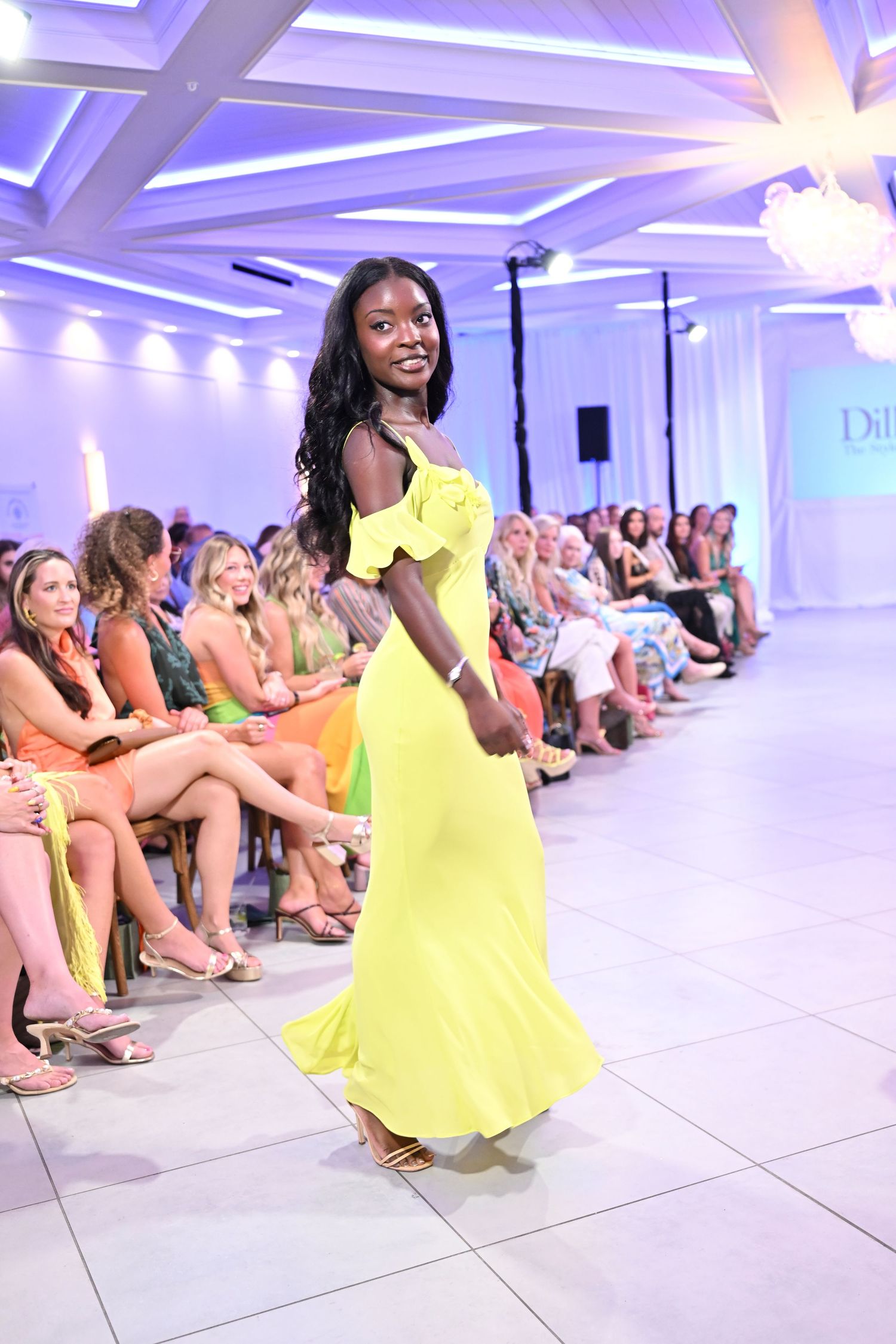Model walks runway in yellow off-shoulder maxi dress at fashion show with seated audience.