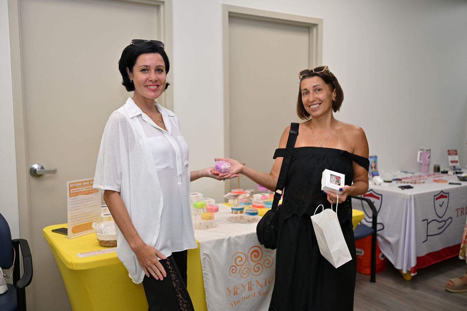Two women smile while exchanging a pink product at an indoor vendor table displaying colorful items.