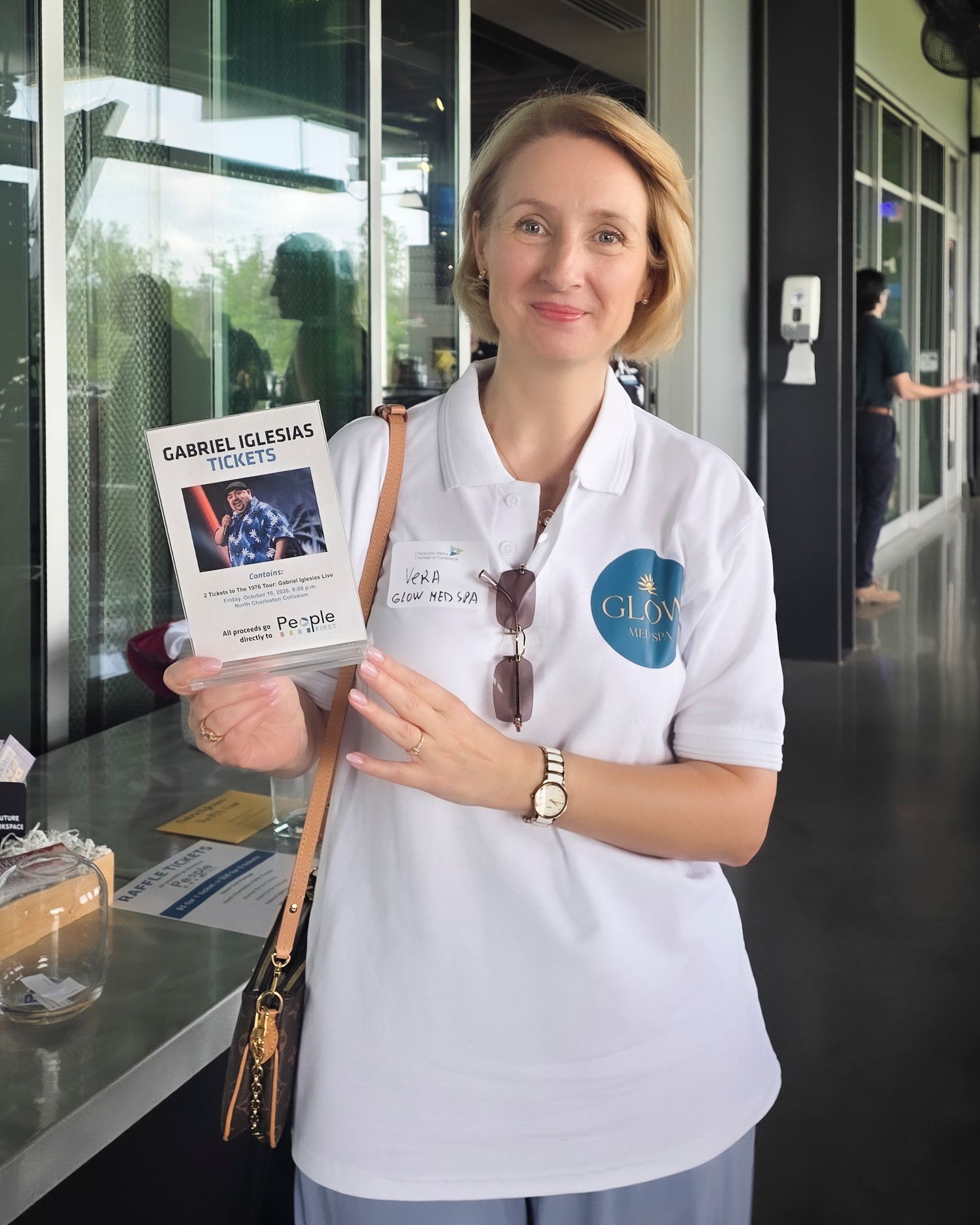 Woman in white polo shirt holding Gabriel Iglesias tickets, wearing a watch and sunglasses, standing indoors.