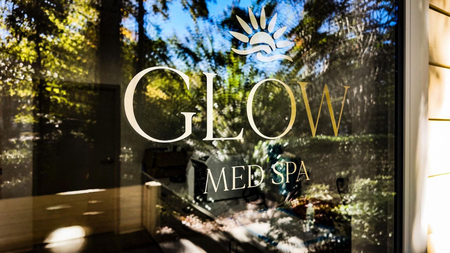 Glow Med Spa glass entrance door with elegant logo surrounded by lush green trees.