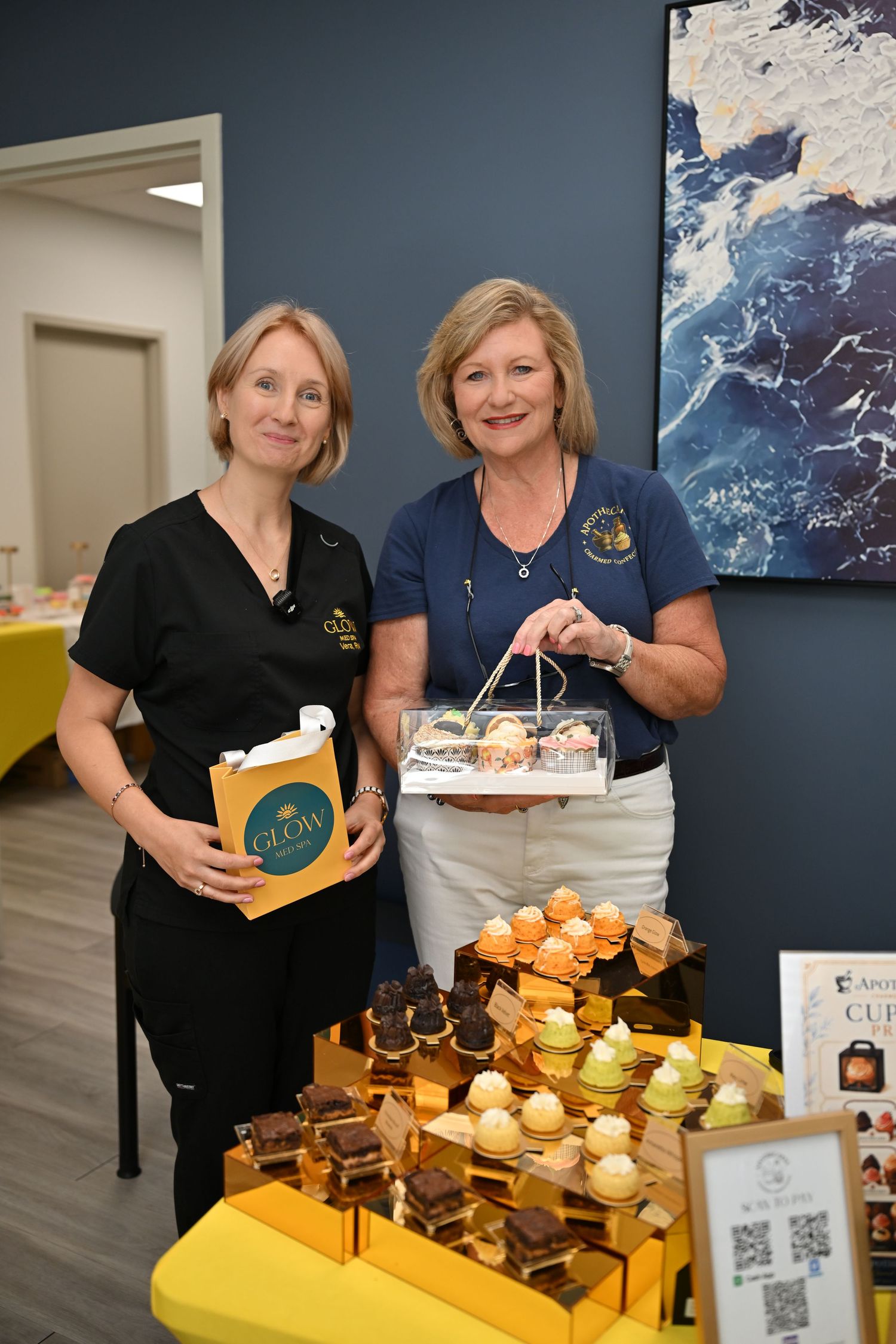 Two women smiling at a dessert table displaying elegant cupcakes and chocolates at a catering or bakery event.