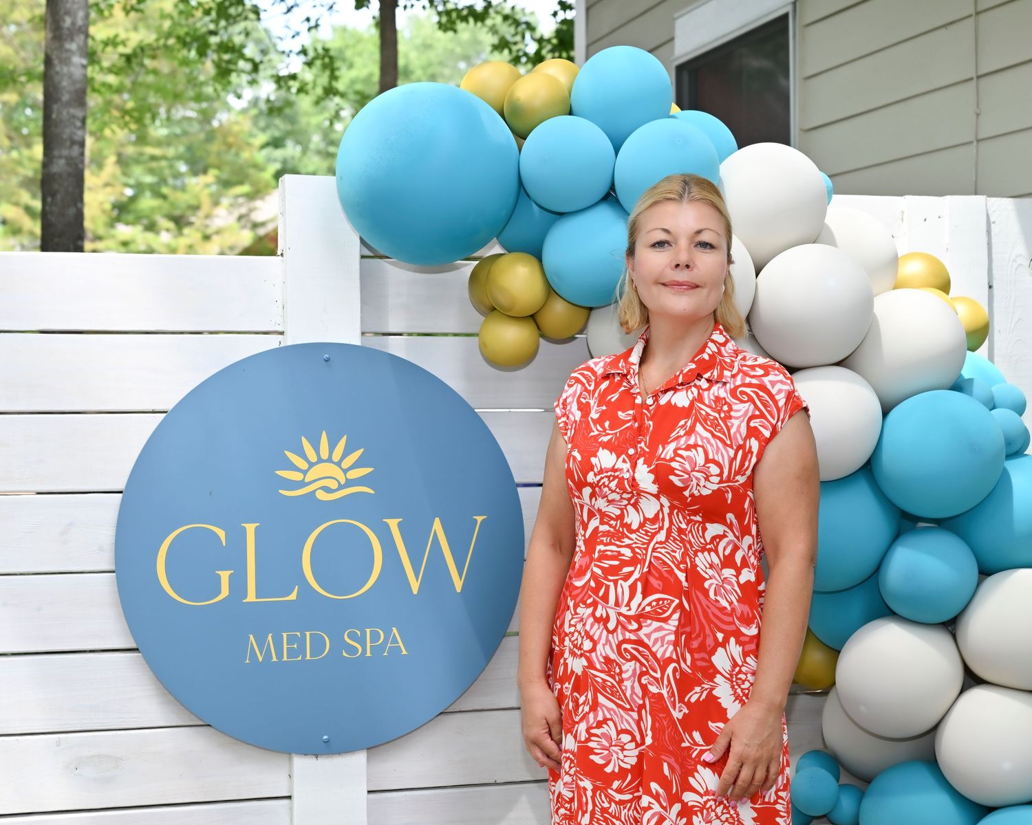 Woman in red floral dress posing beside Glow Med Spa sign with teal and gold balloon arch backdrop.