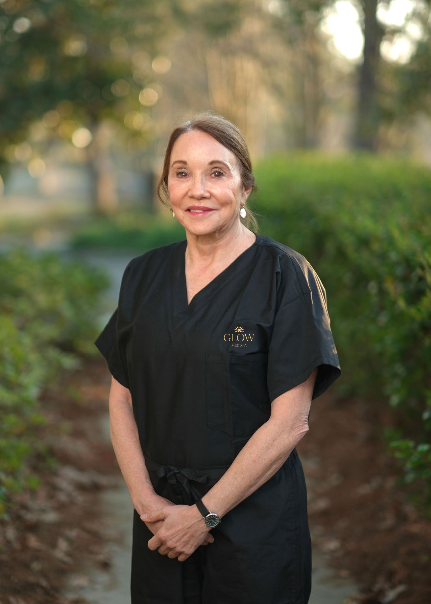 A professional in black Glow medical scrubs stands confidently outdoors in a serene garden pathway.