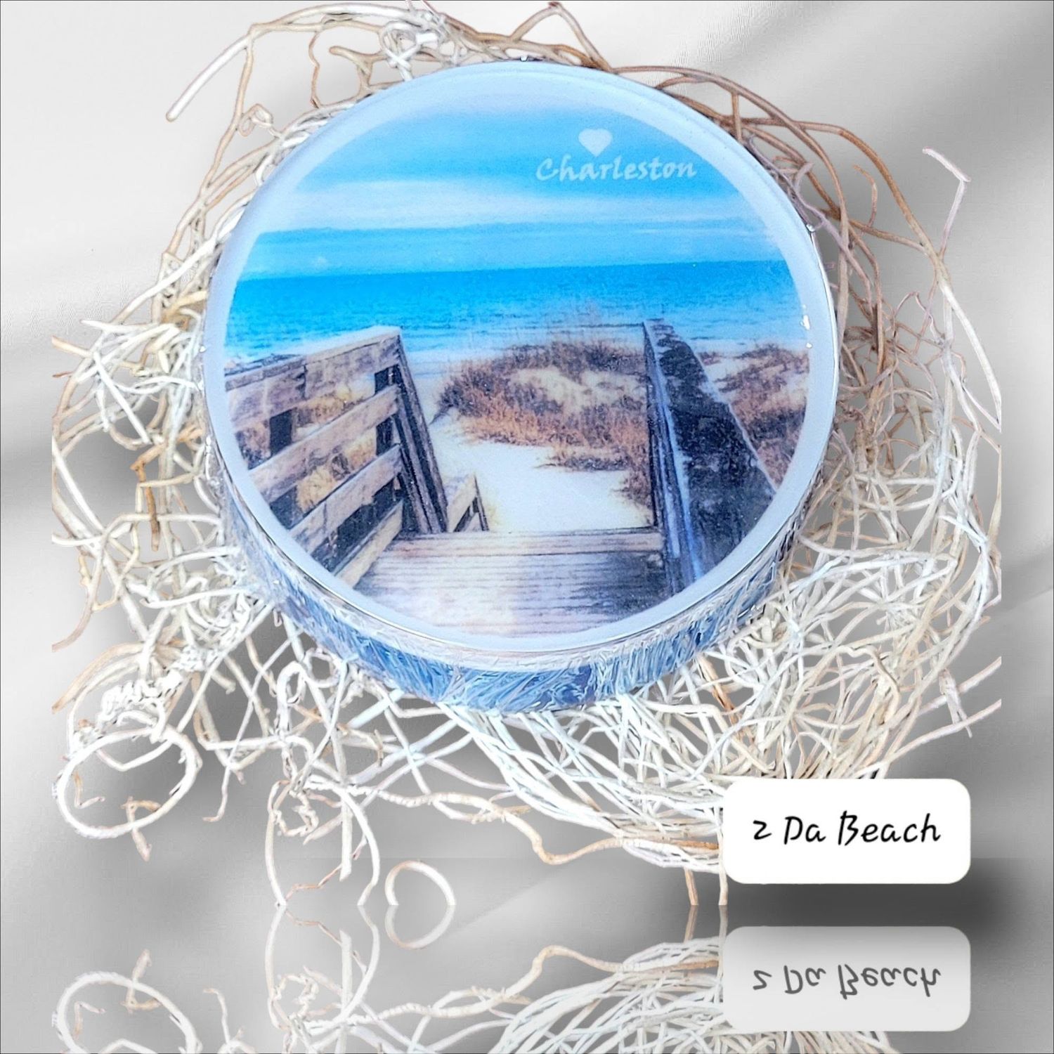 Round Charleston beach scene candle tin with boardwalk and ocean view, nestled in white raffia gift packaging.