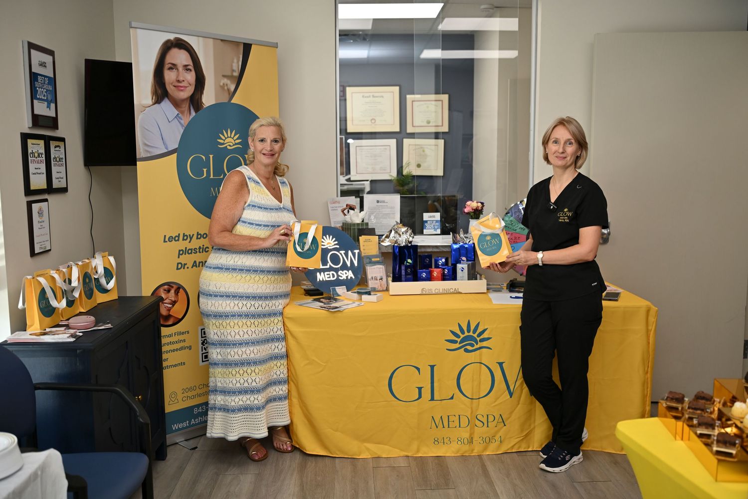 Two women stand at a GLOW Med Spa branded table display with products and promotional banners at a business event.