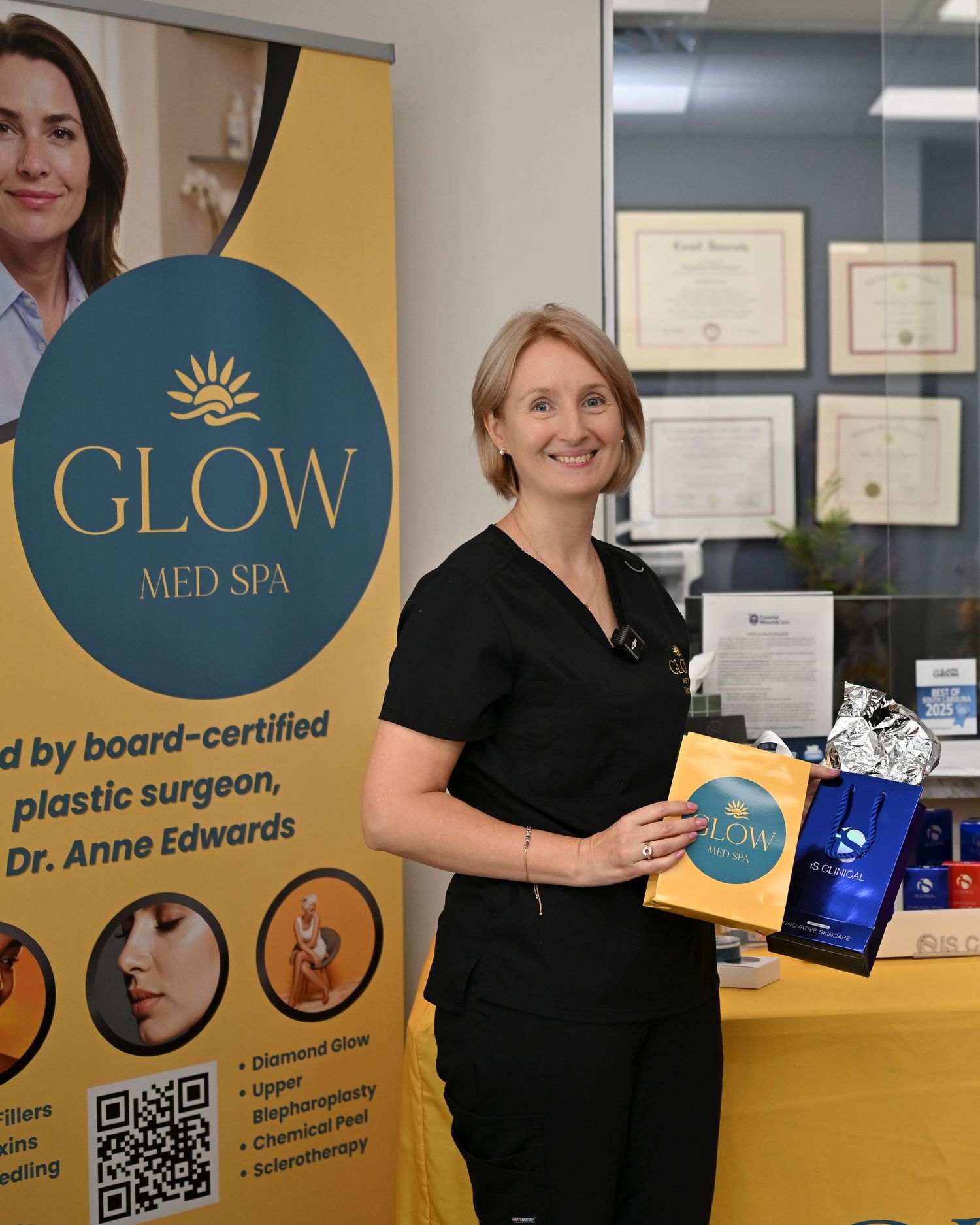 Dr. Anne Edwards smiles holding Glow Med Spa products beside a promotional banner at her board-certified plastic surgery spa.