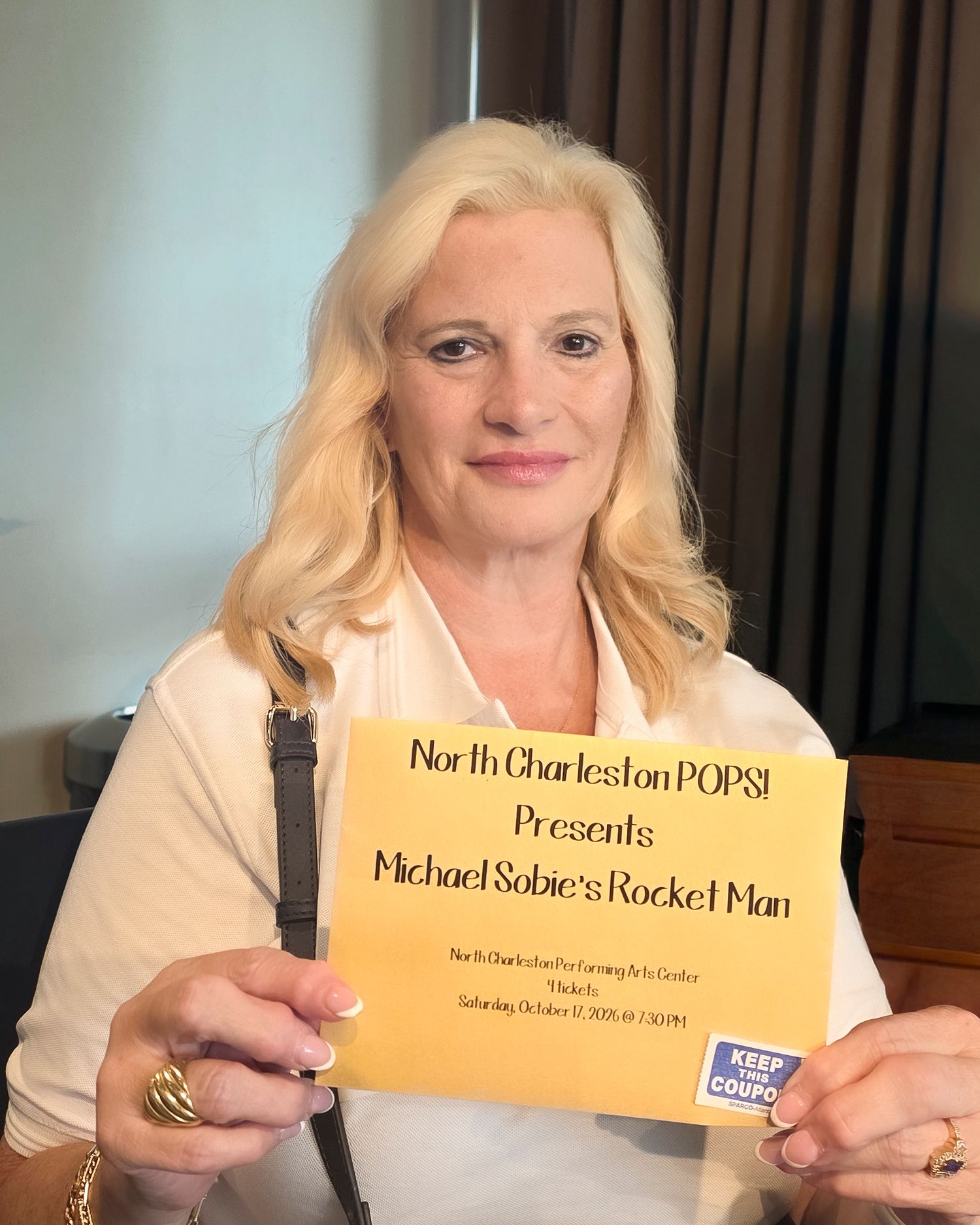 Woman holding a yellow North Charleston POPS presents Michael Sobie's Rocket Man event flyer at a venue.