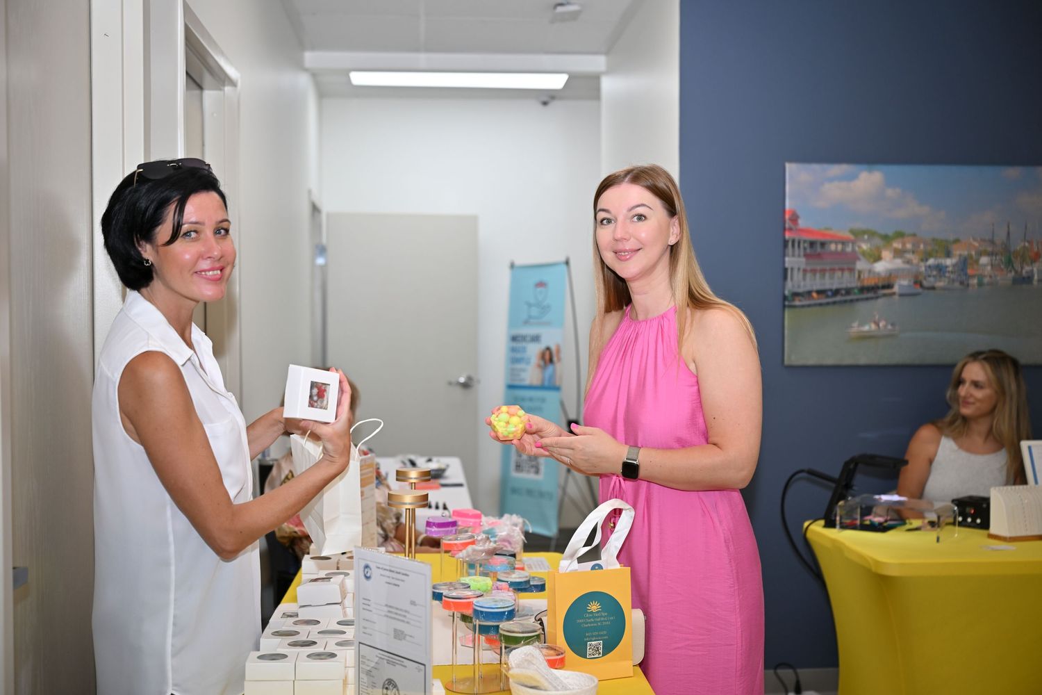 Two women smiling at a vendor table display with products, in a bright modern indoor event space.