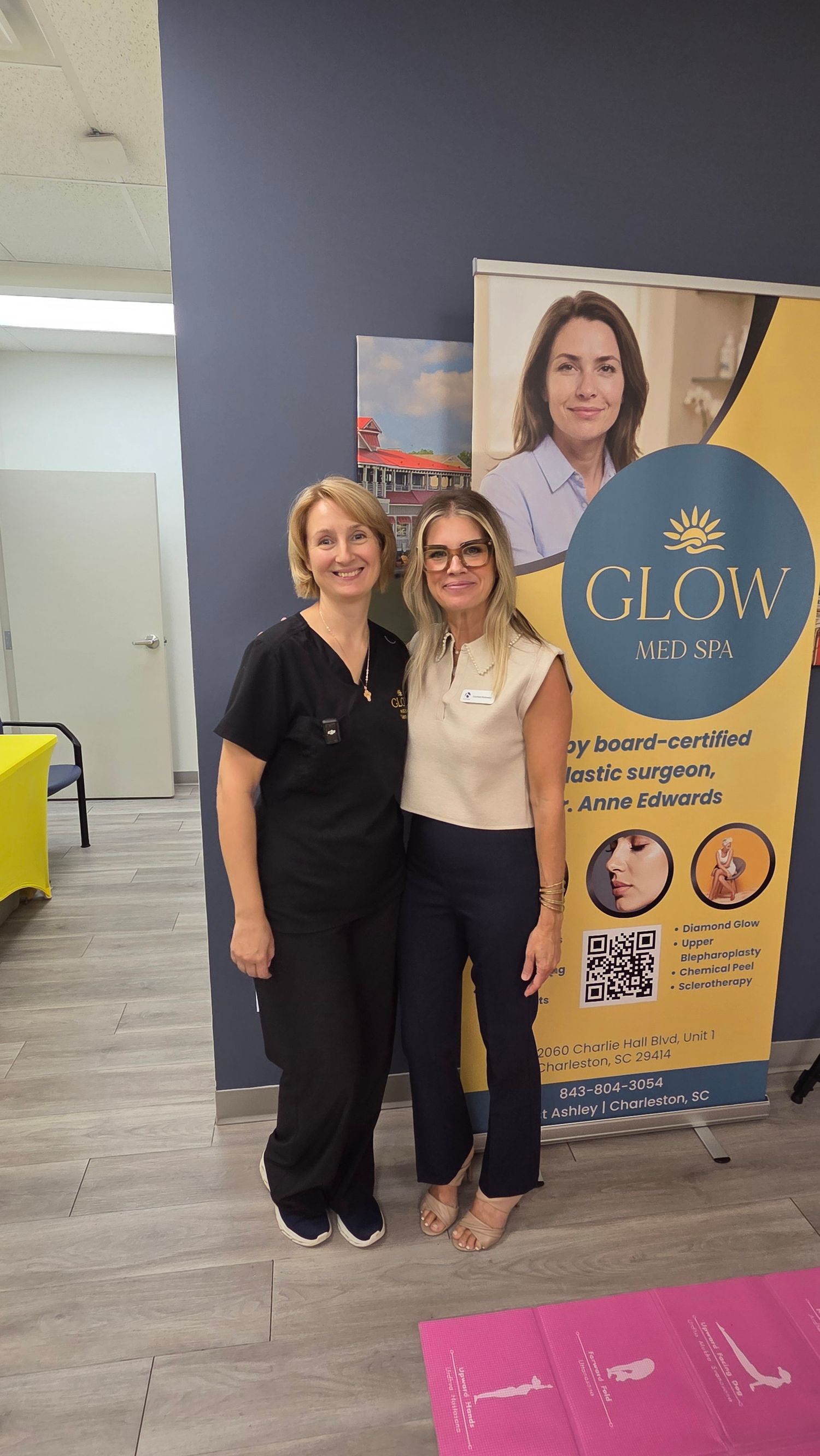 Two women smiling together in front of a Glow Med Spa banner in Charleston, SC.
