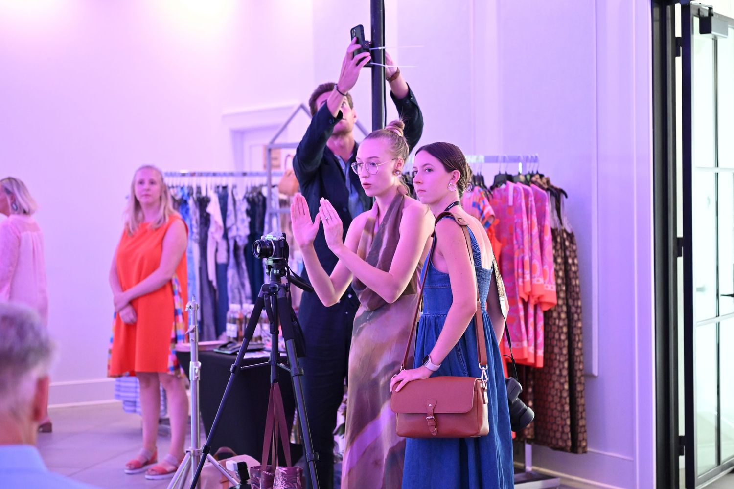 Dancers in blue gowns perform at a fashion event with clothing racks and cameras in a pink-lit showroom.