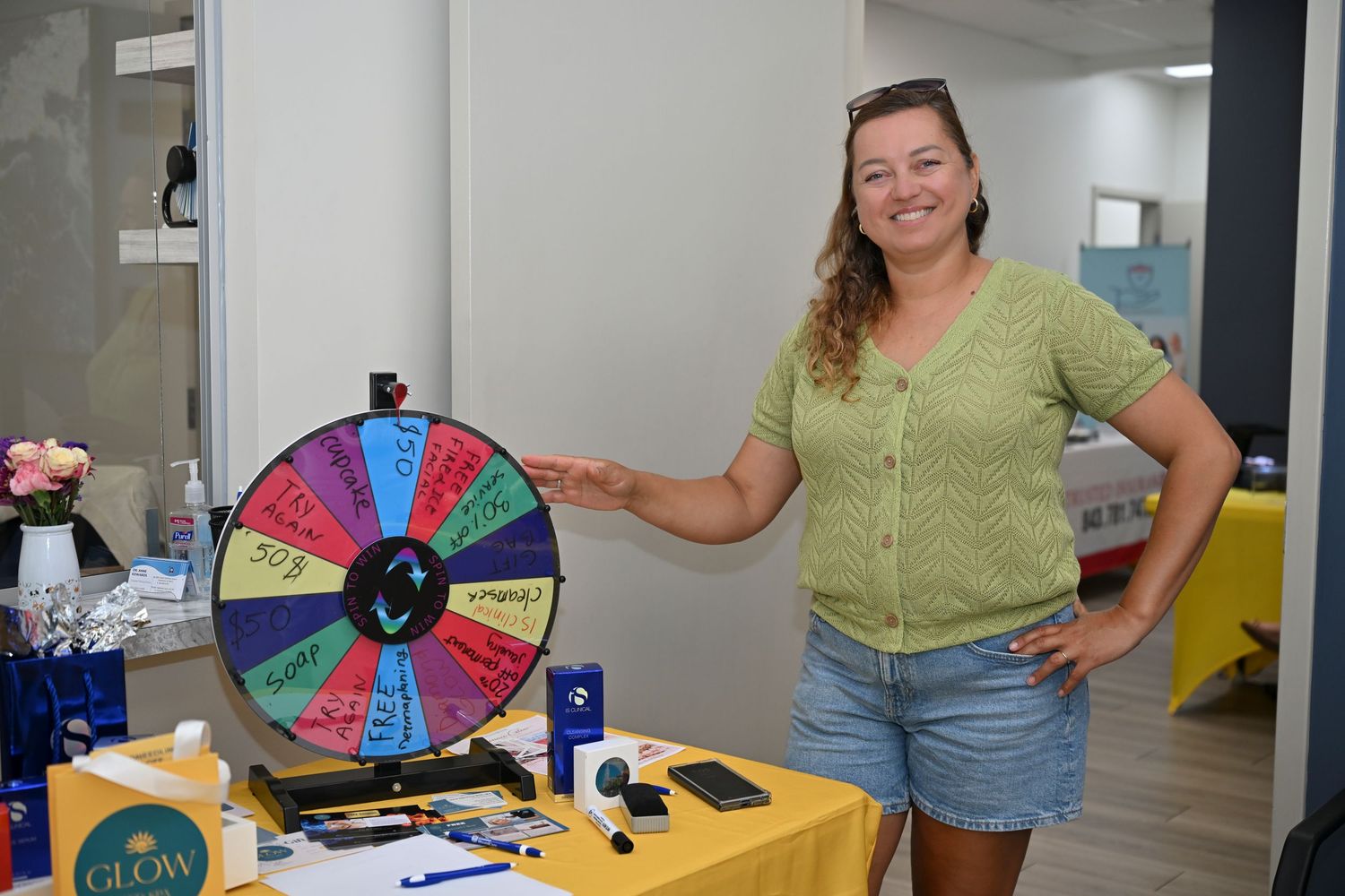 Woman smiling beside a colorful prize spin wheel at an indoor event booth with promotional items on table.