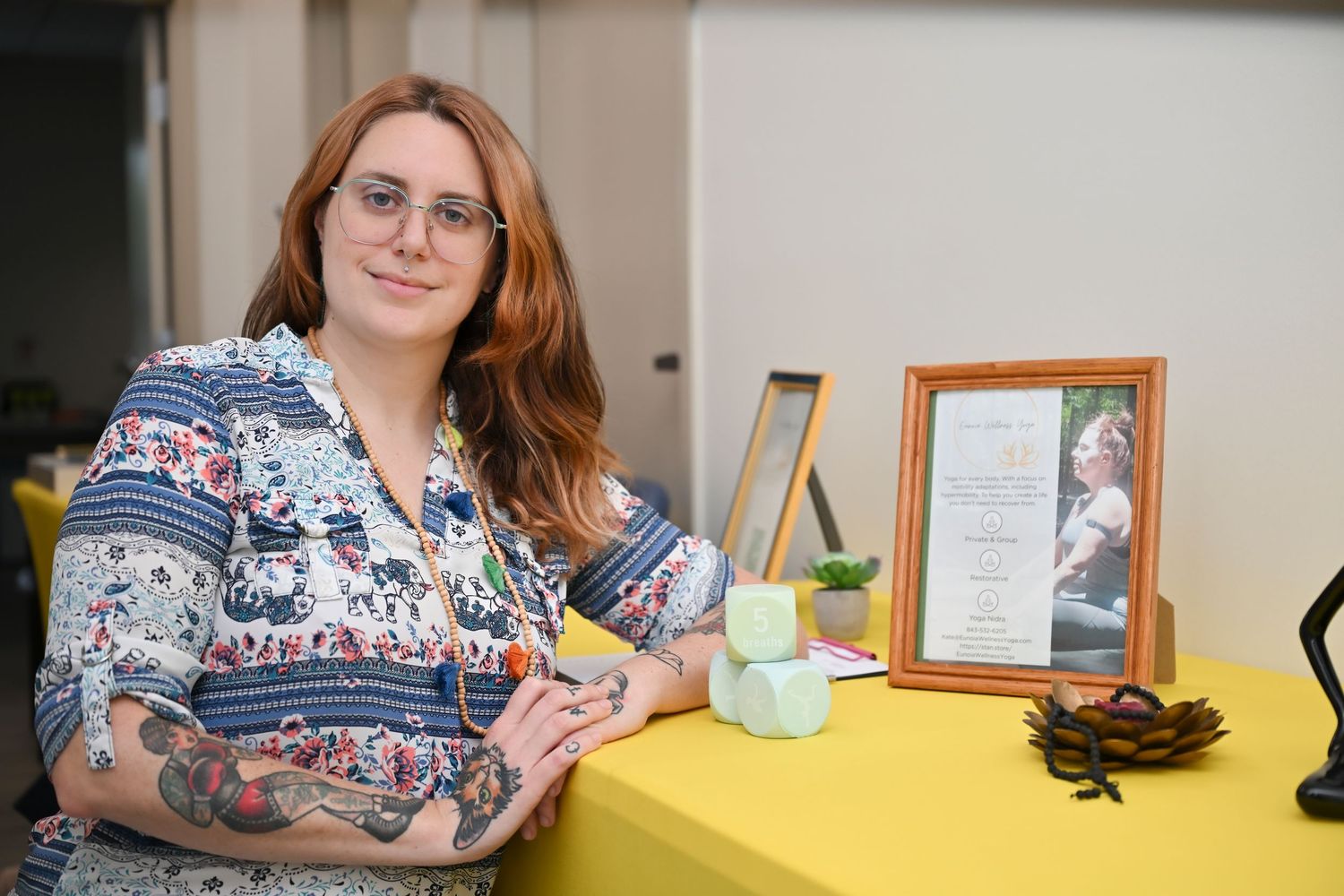 Woman with red hair and tattoos poses beside a framed photo and decorative items on a yellow table.