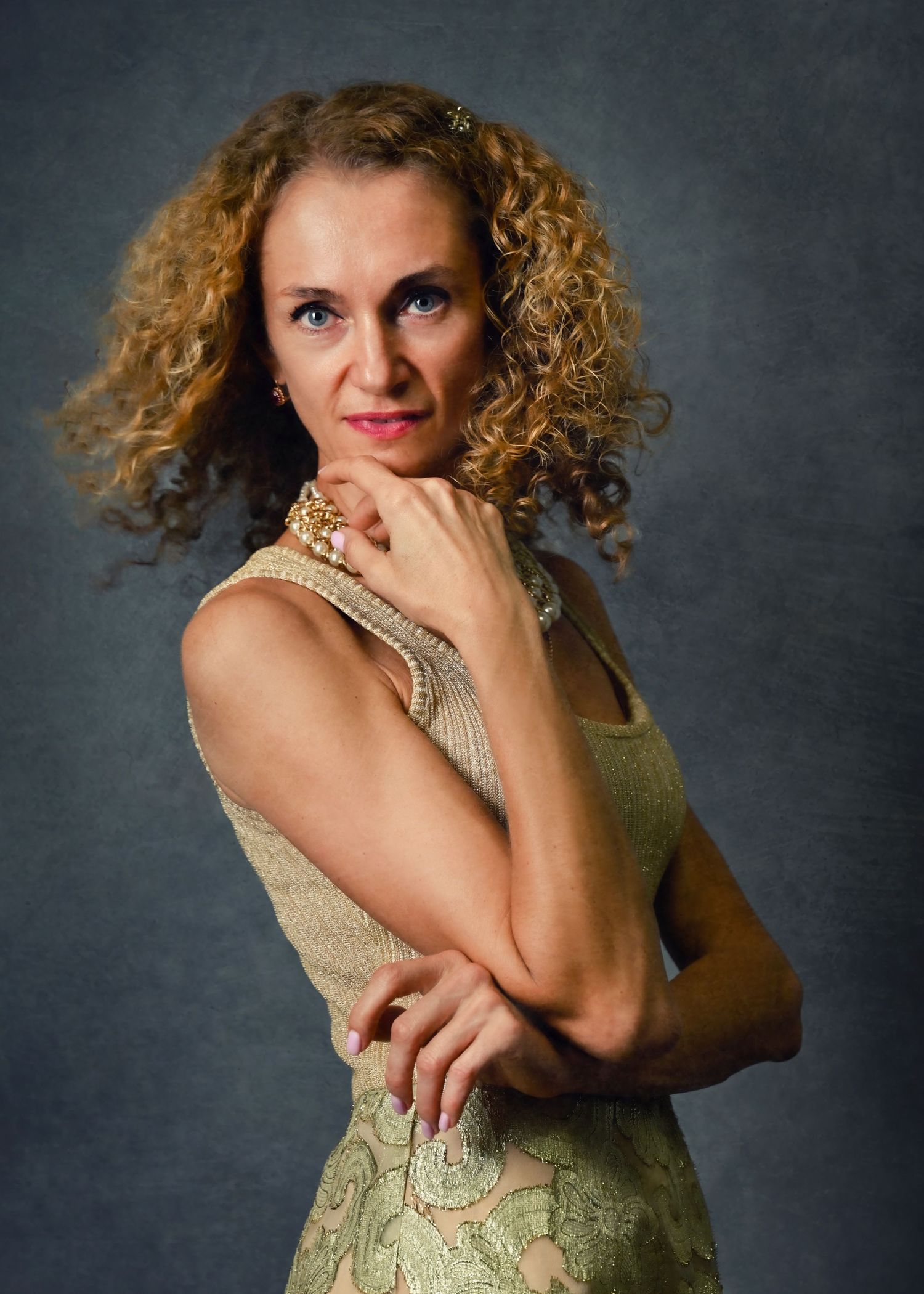 A curly-haired woman in a gold lace dress poses confidently against a gray studio background.