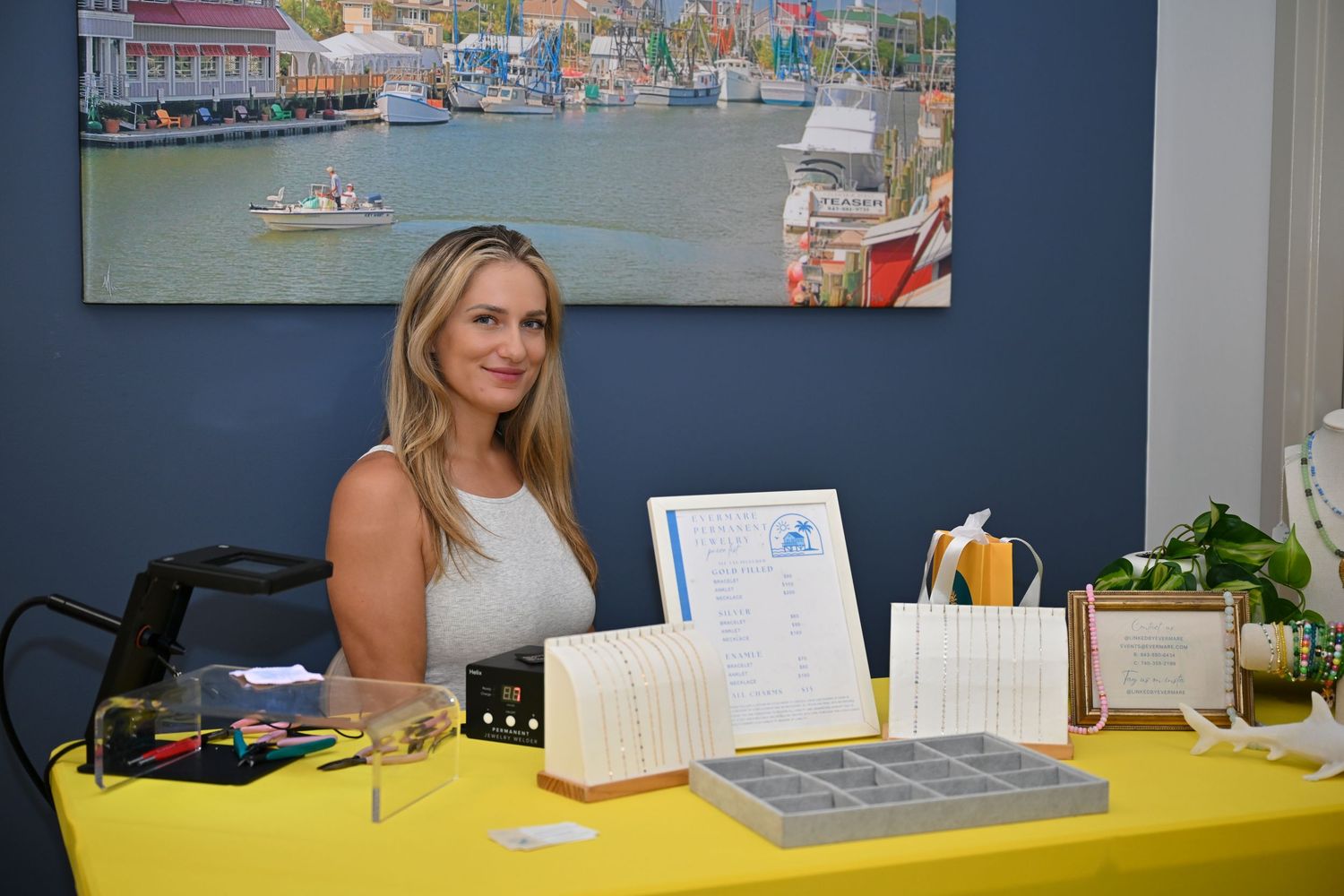 Smiling woman at a yellow-draped vendor table displaying products indoors with a harbor painting backdrop.