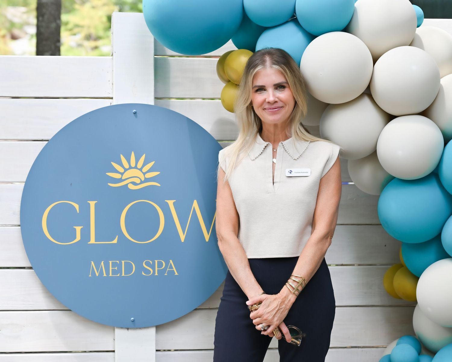 Woman posing in front of Glow Med Spa sign with teal and gold balloon arch backdrop.