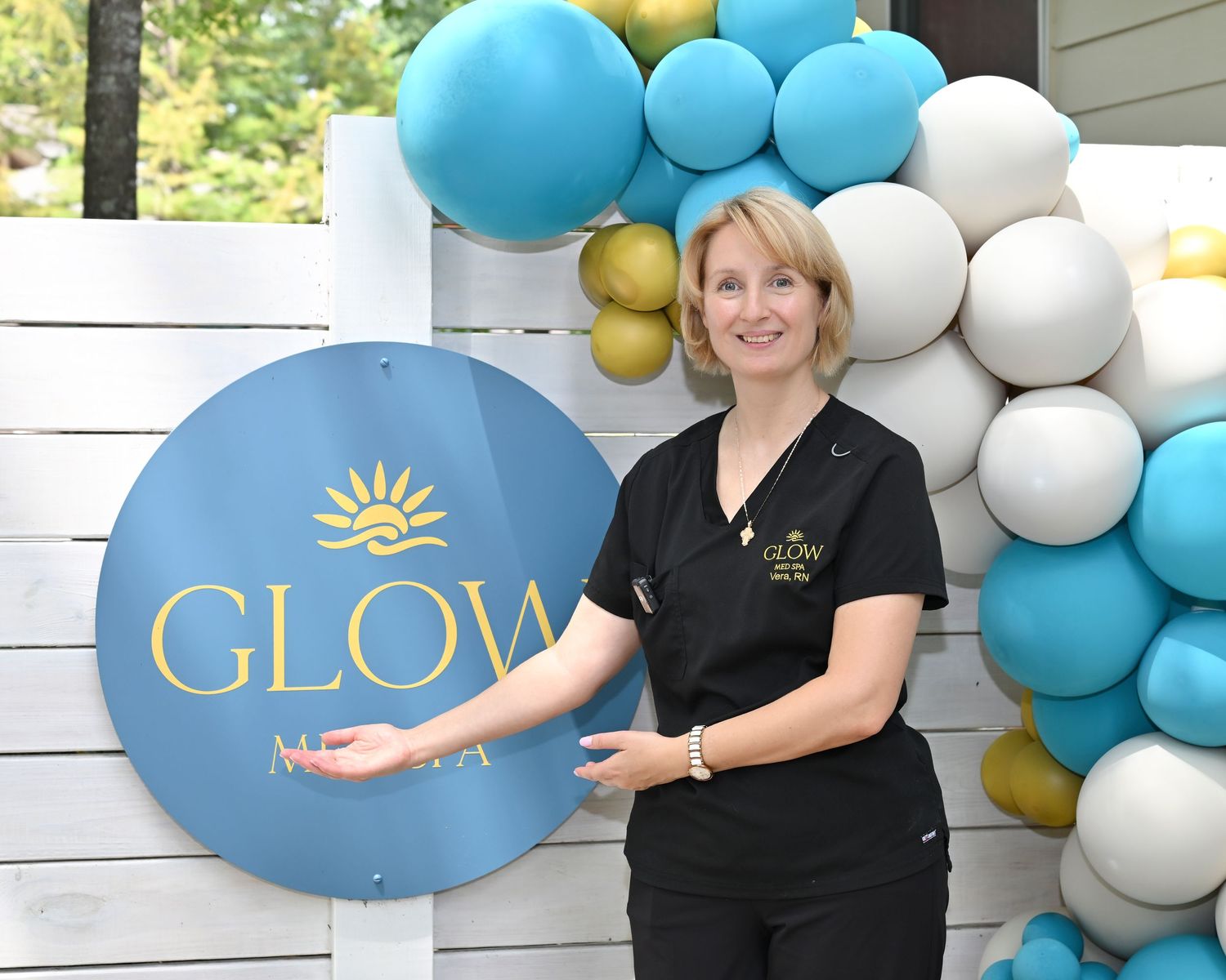 Woman in black uniform smiling beside a round Glow Med Spa sign with teal and gold balloon arch.