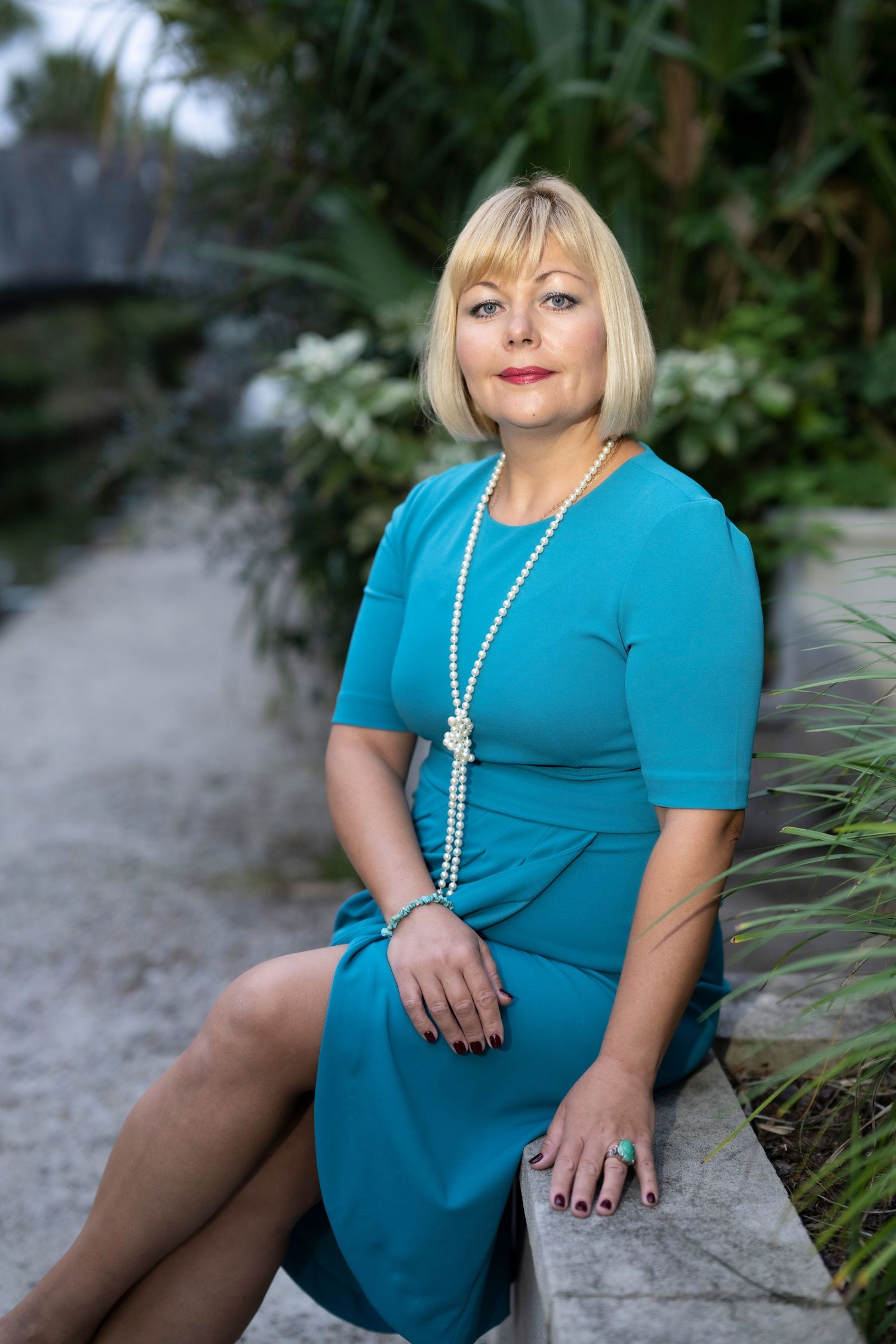 A blonde woman in a teal dress and pearl necklace sits outdoors in a lush garden setting.