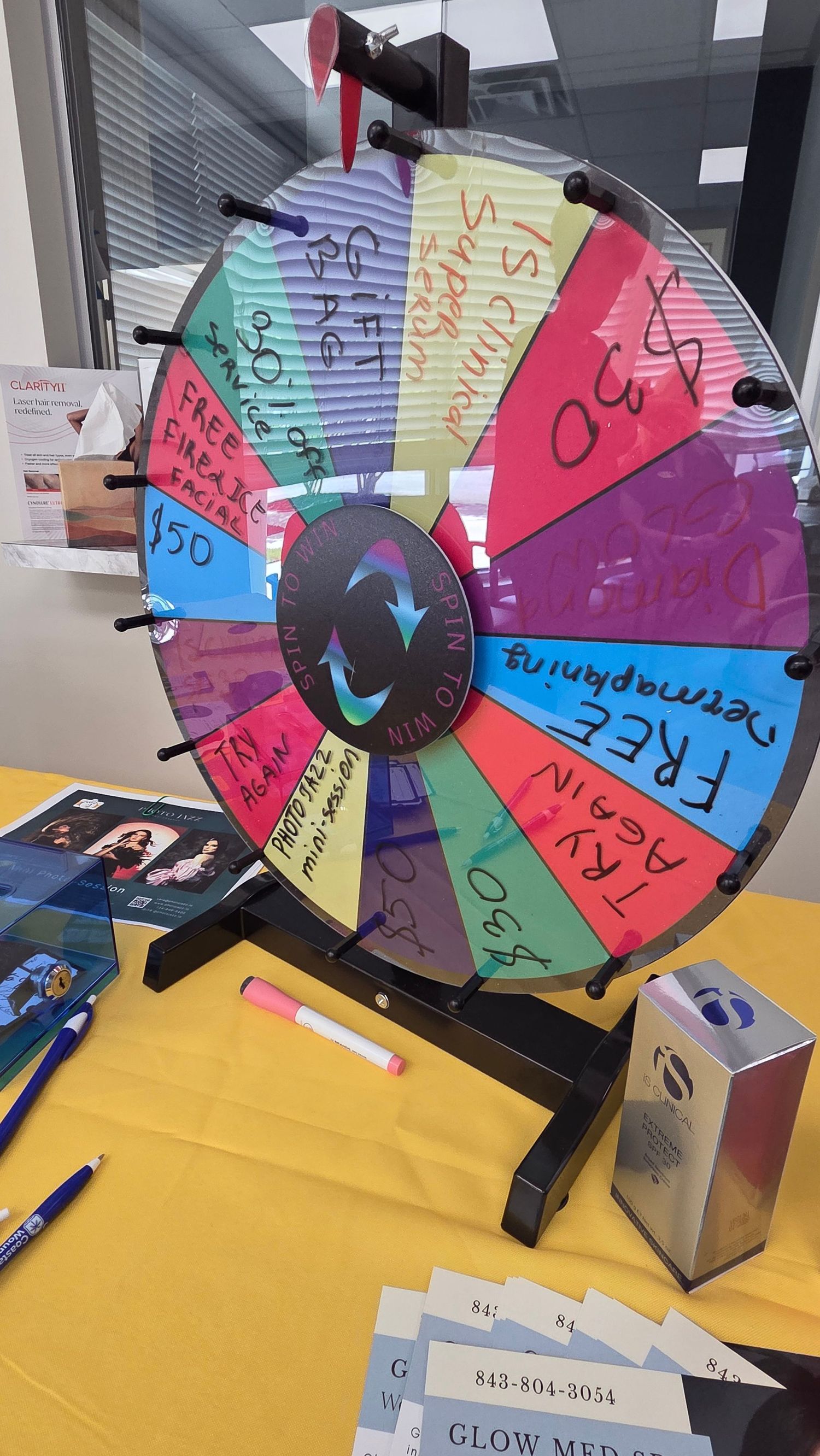 Colorful prize spinning wheel on a yellow table at Glow Med Spa promotional event with gift cards and pens.