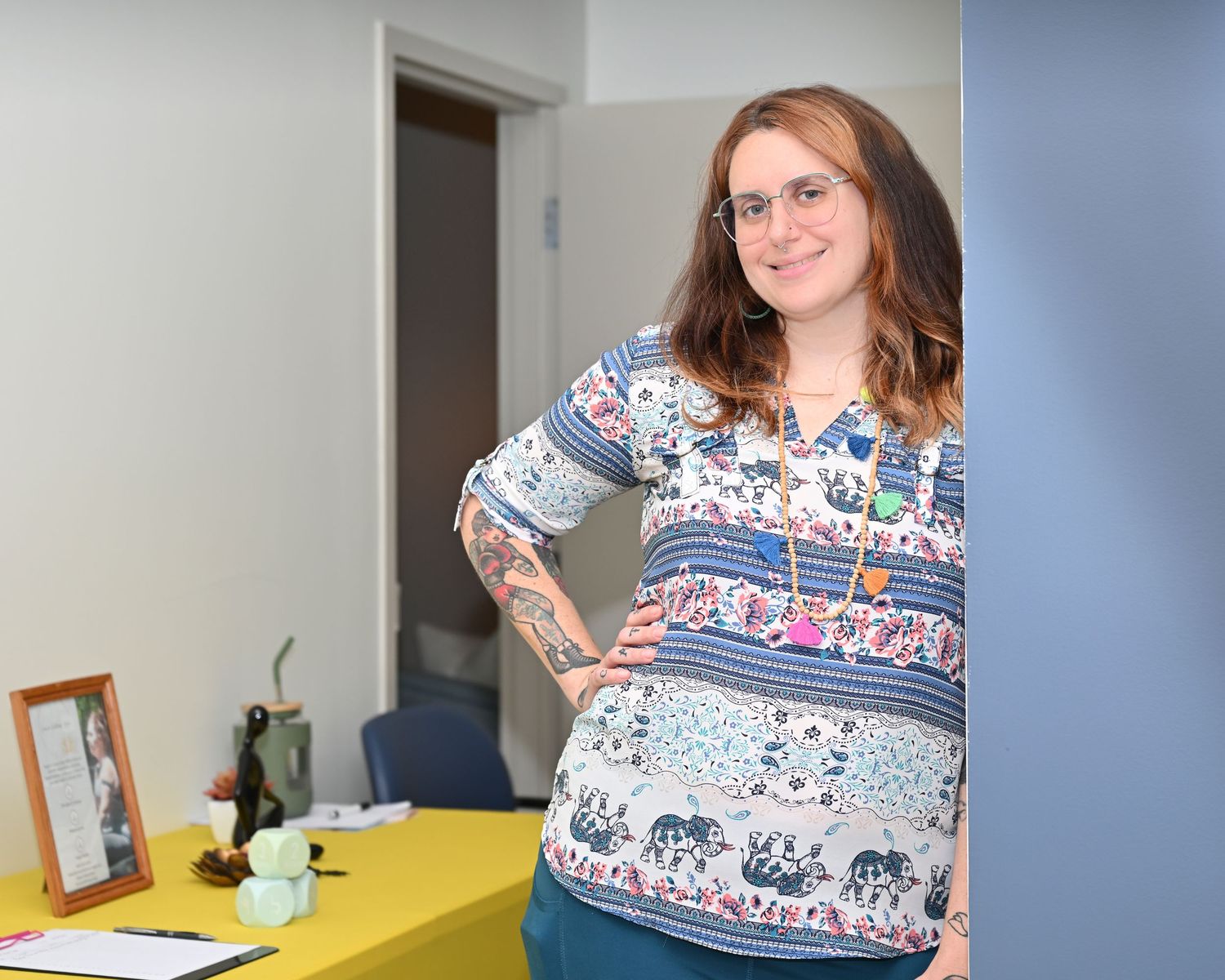 Woman with tattoos in floral blouse smiling, posing in an office setting with a yellow desk and framed photo.