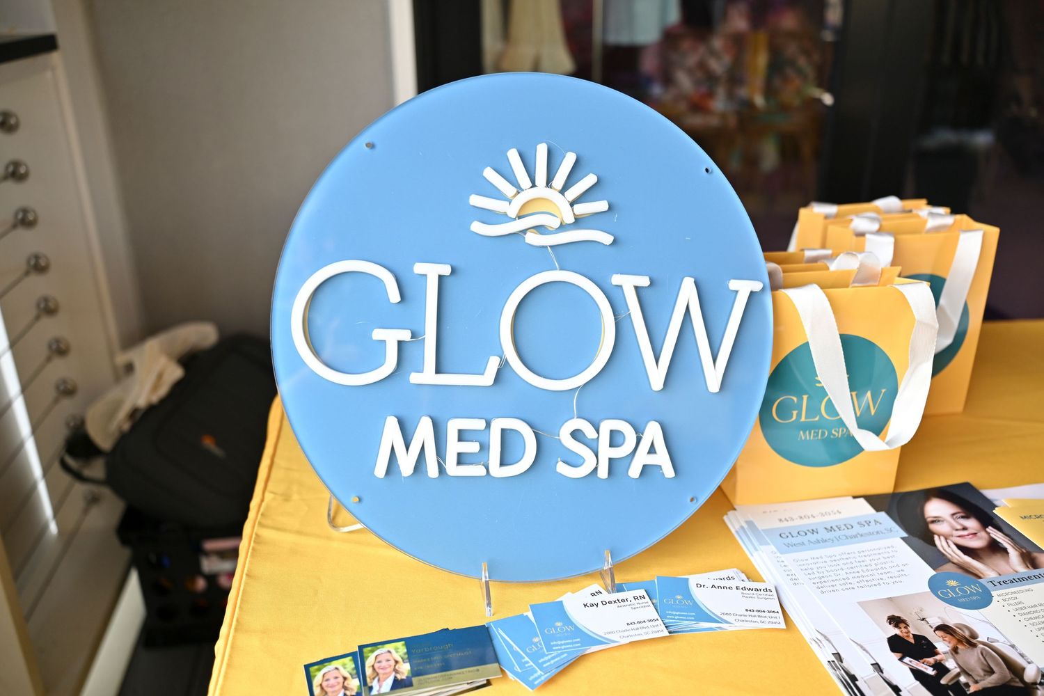 Glow Med Spa blue circular sign displayed on a yellow table with brochures and tote bags.