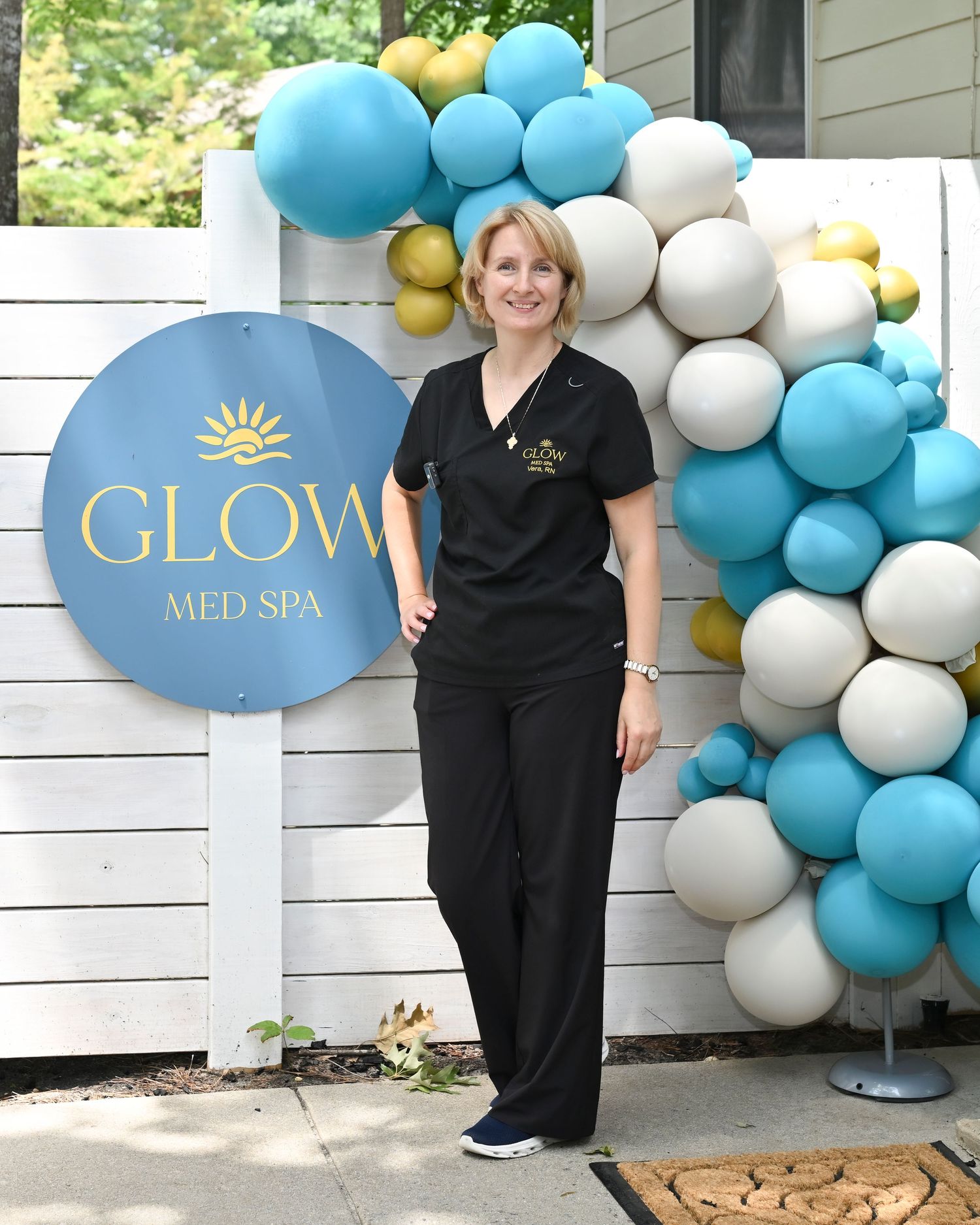 Woman in black scrubs poses beside Glow Med Spa sign with teal, gold, and white balloon arch outdoors.