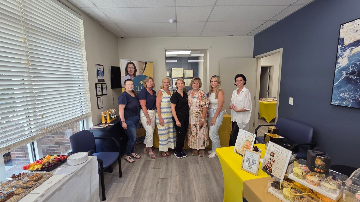 Group of eight women smiling together in a bright office reception area with yellow accents and snack display.