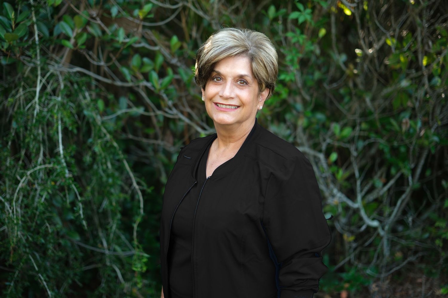 Smiling woman in black top standing outdoors among green foliage.