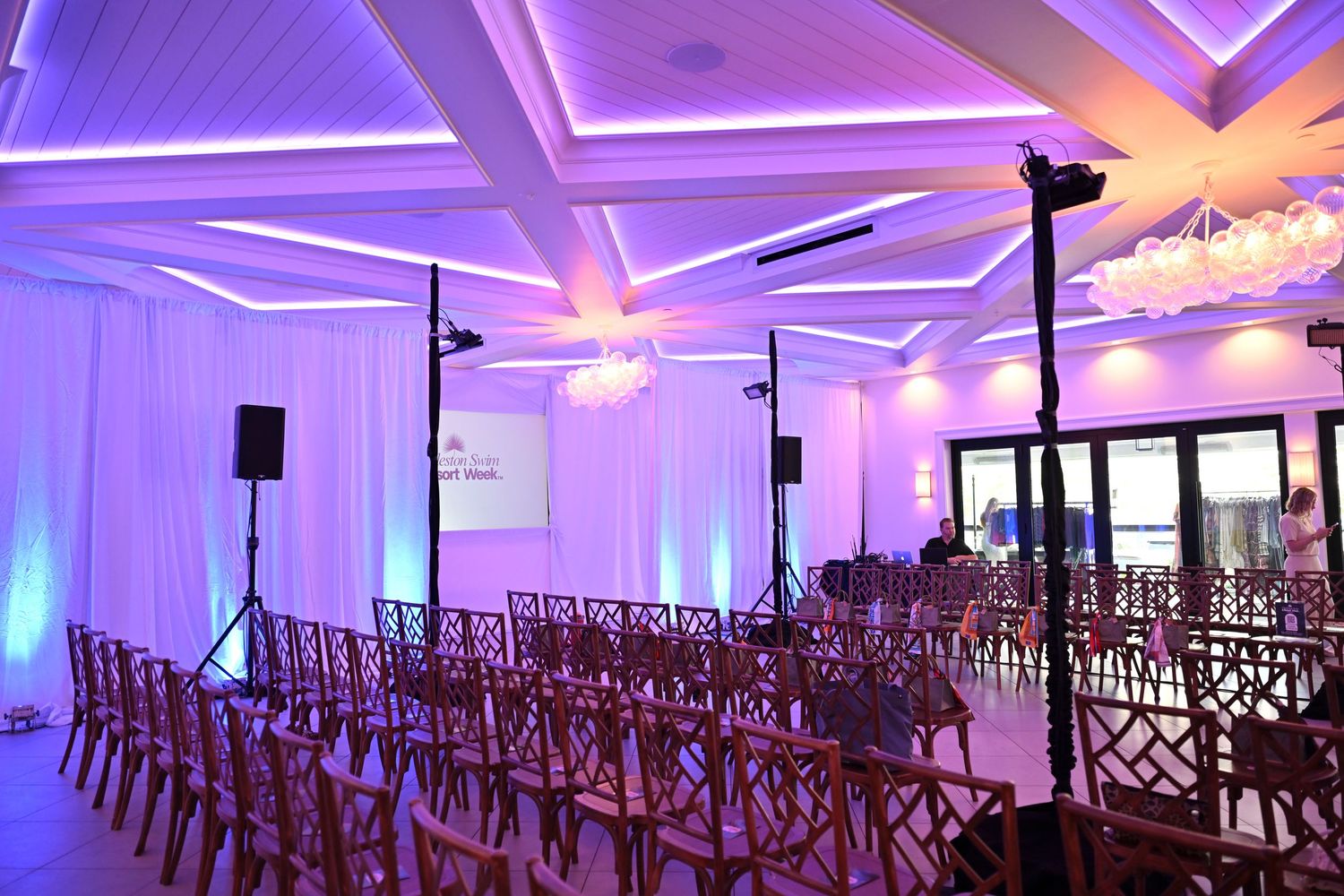 Elegant event venue with purple uplighting, rows of wooden chairs, and white draping for a wedding ceremony setup.