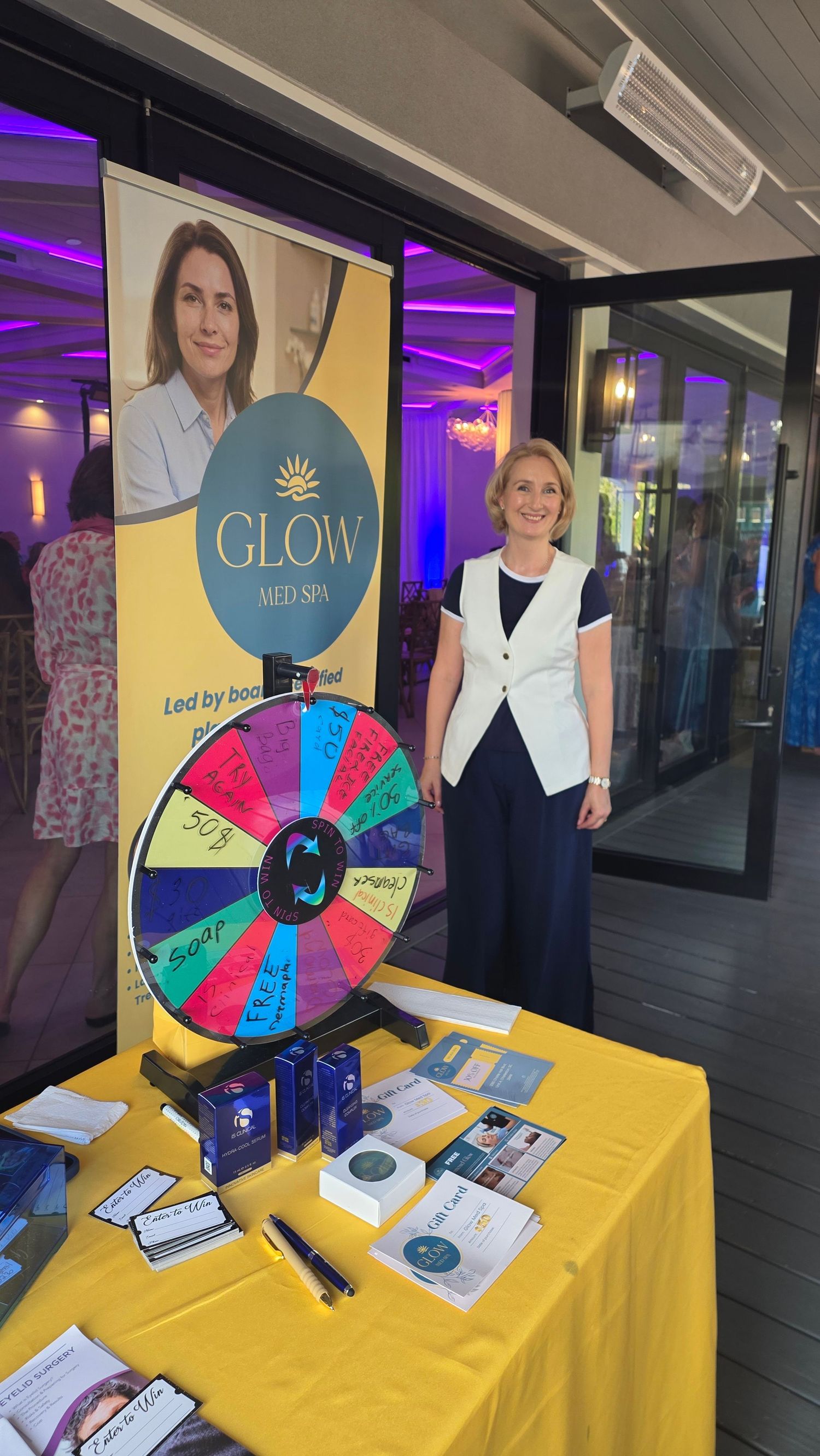 Woman standing beside a prize wheel at a Glow Med Spa promotional booth with branded banner and yellow tablecloth.
