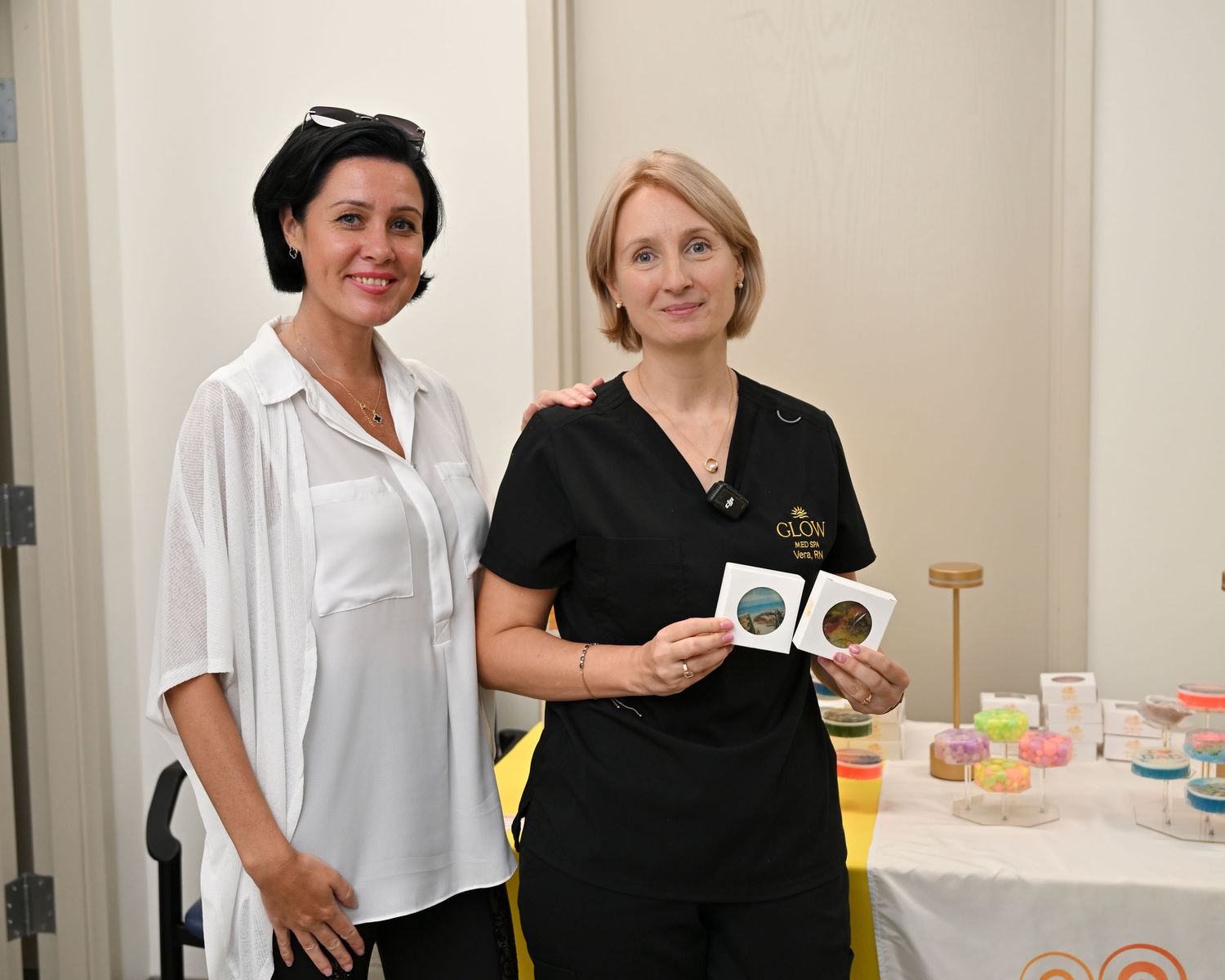 Two women posing together at a beauty product display table, one holding cosmetic compacts in a bright indoor setting.