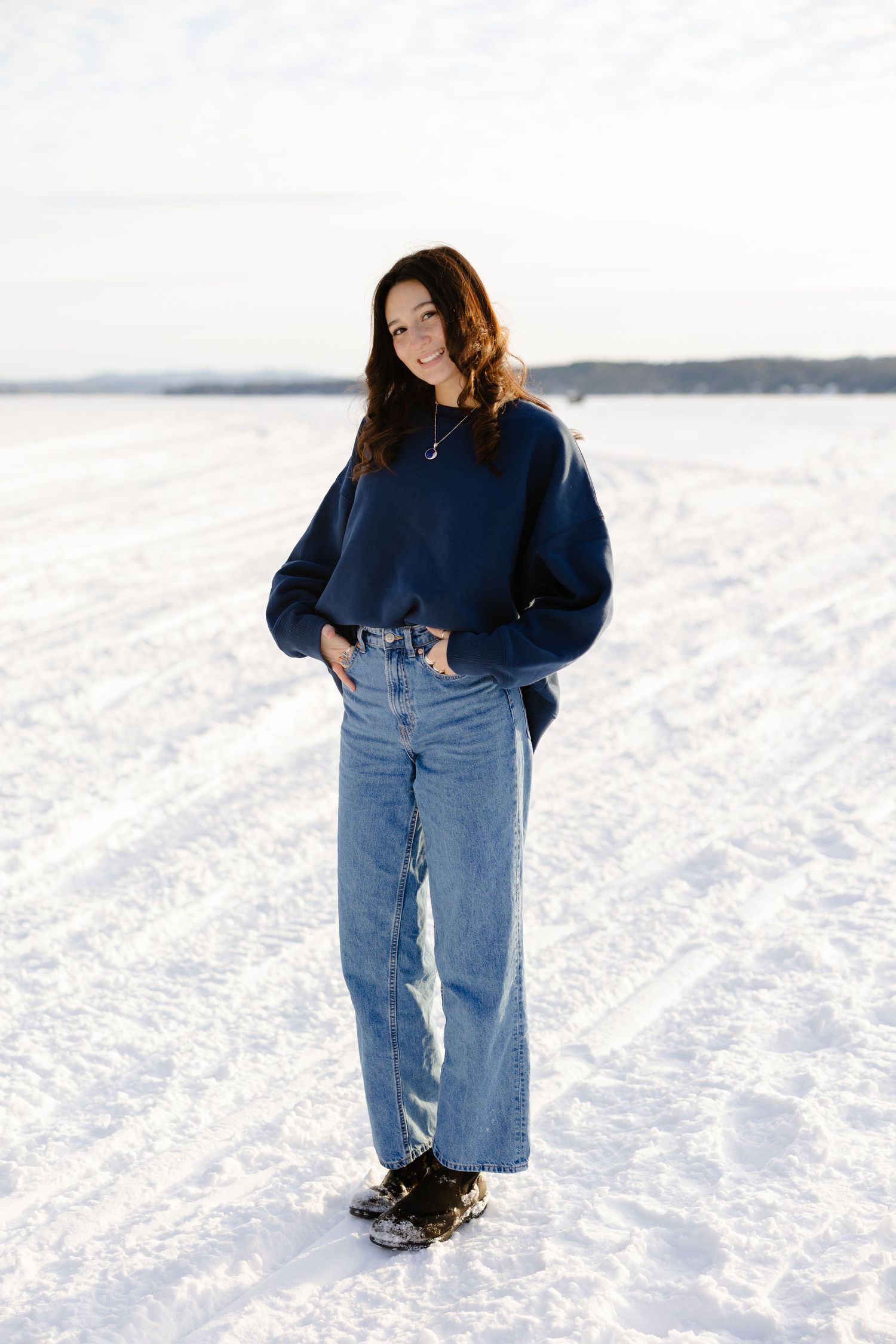 Young woman in navy sweatshirt and wide-leg jeans standing on snowy landscape in winter outdoor setting.