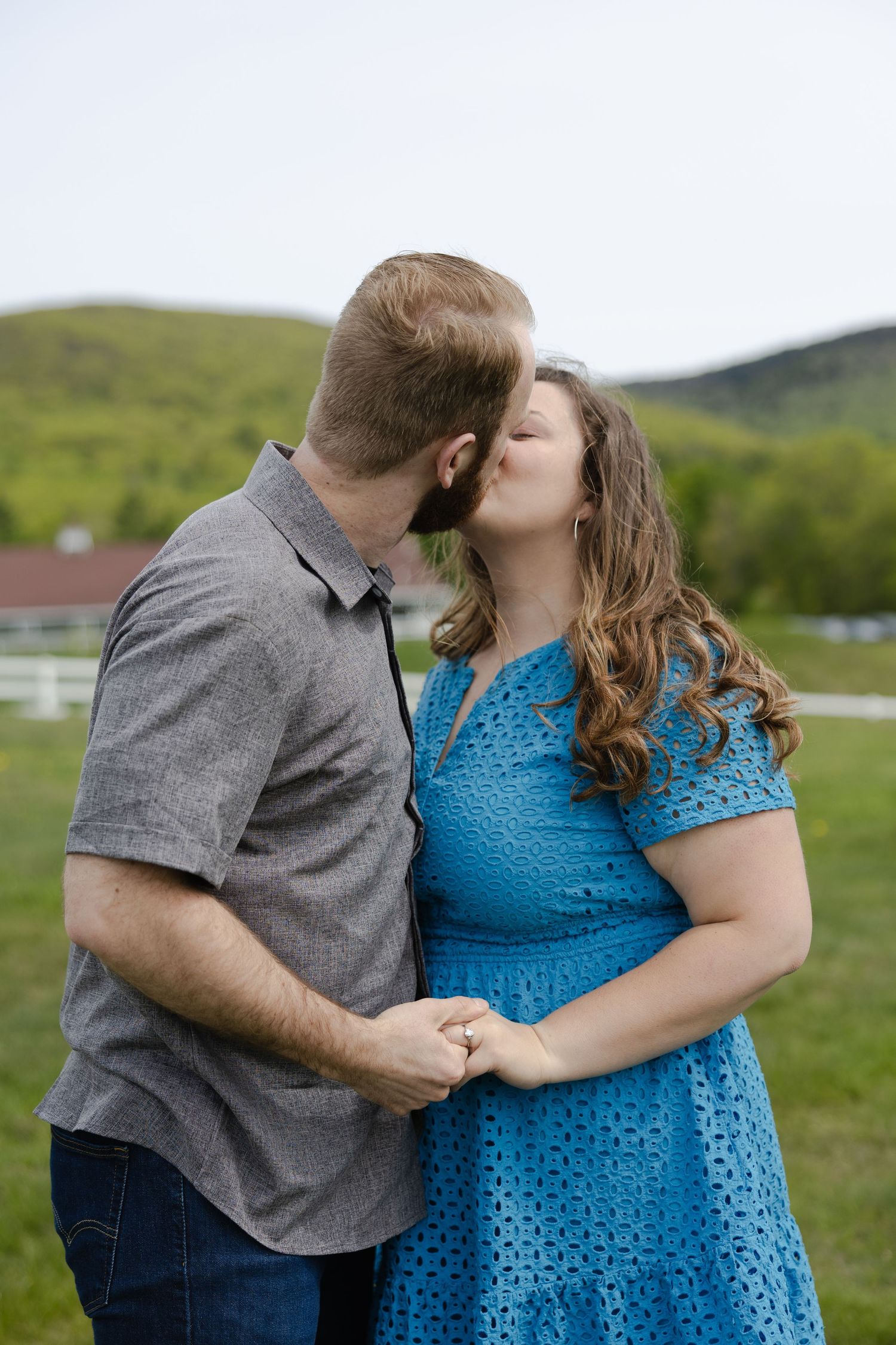 Couple sharing a romantic kiss outdoors, woman in blue lace dress holding hands with man in gray shirt, green hills behind.