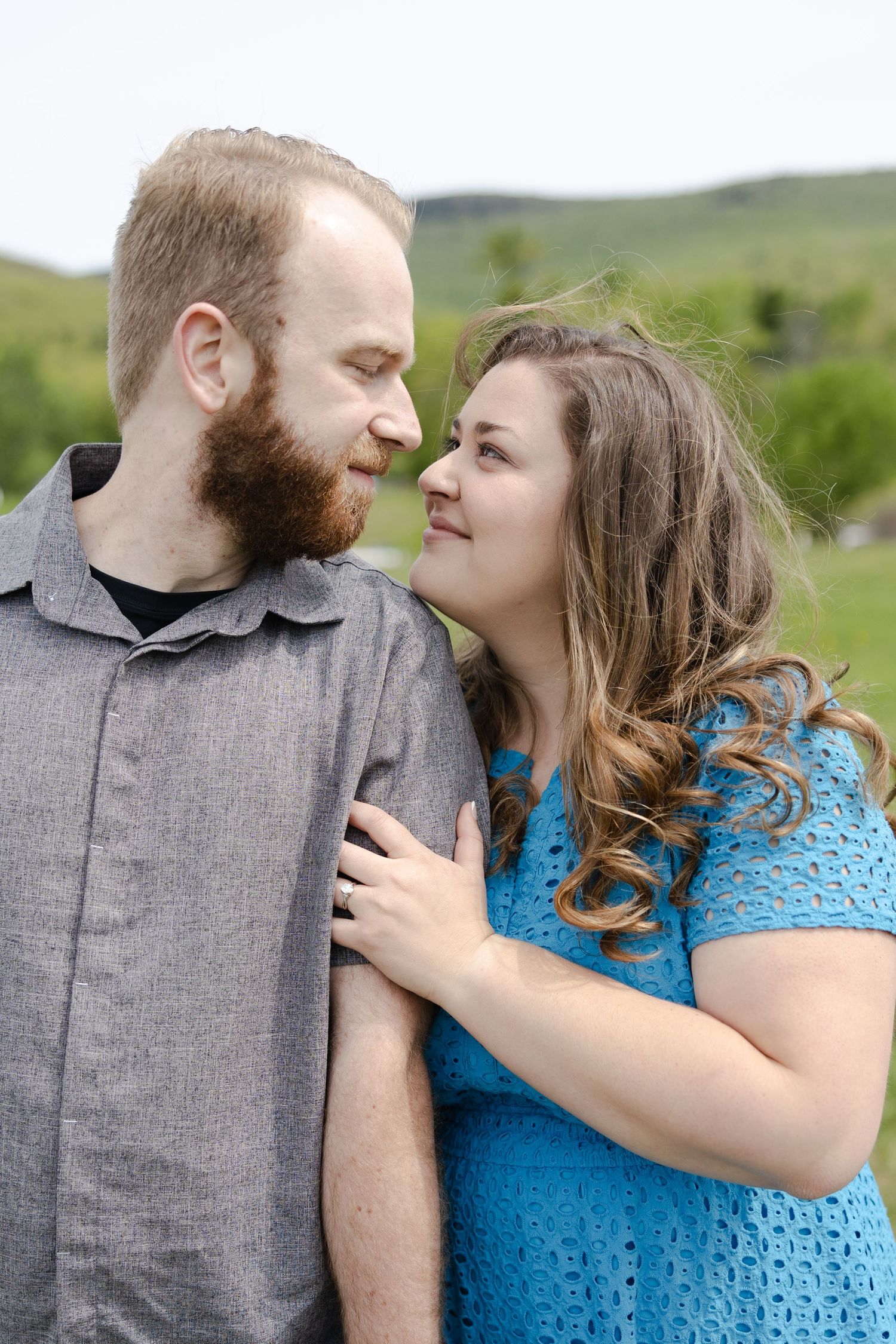 A bearded man and smiling woman in blue share a romantic moment outdoors with green hills behind them.