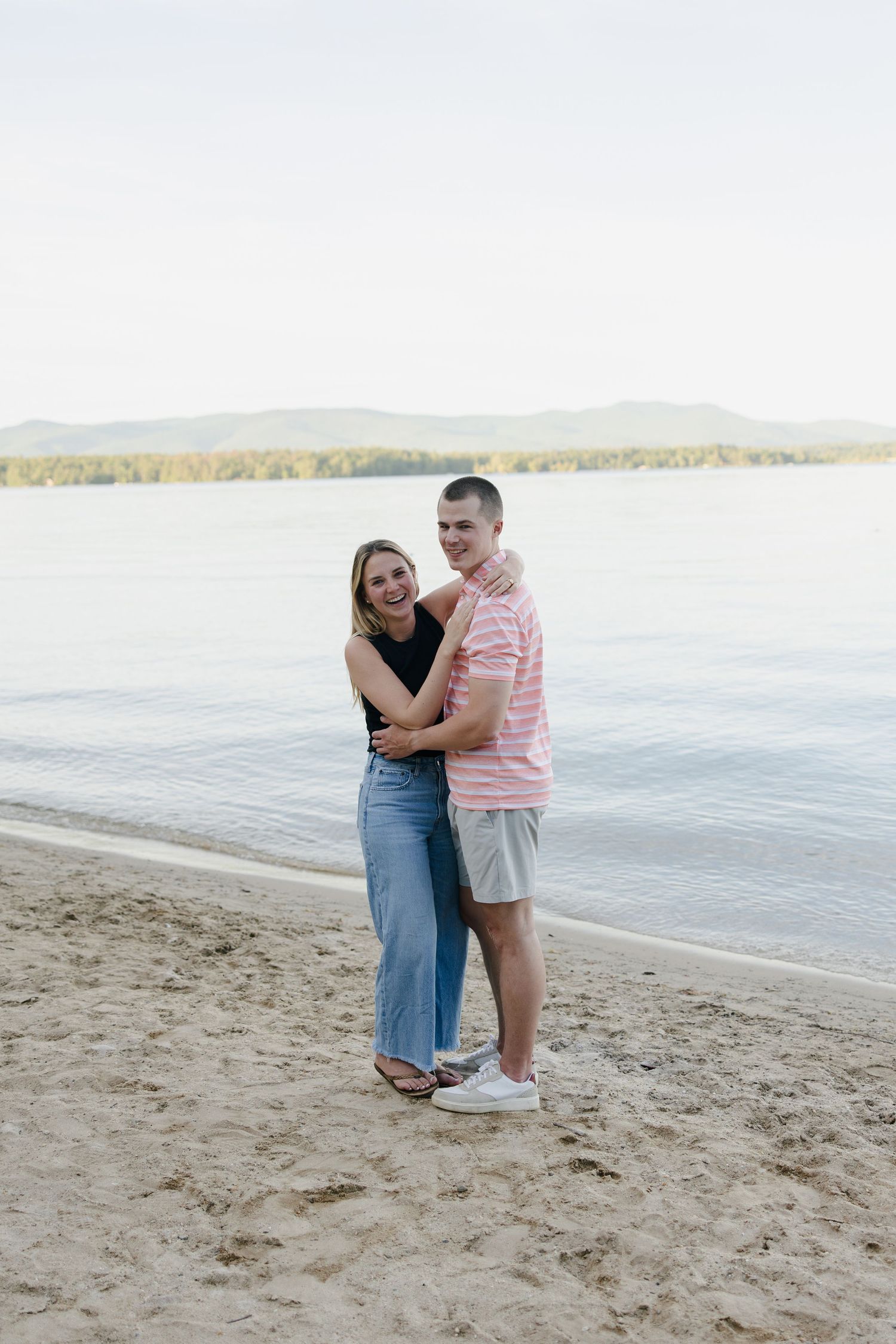A couple embraces and smiles on a sandy beach with calm water and mountains in the background.