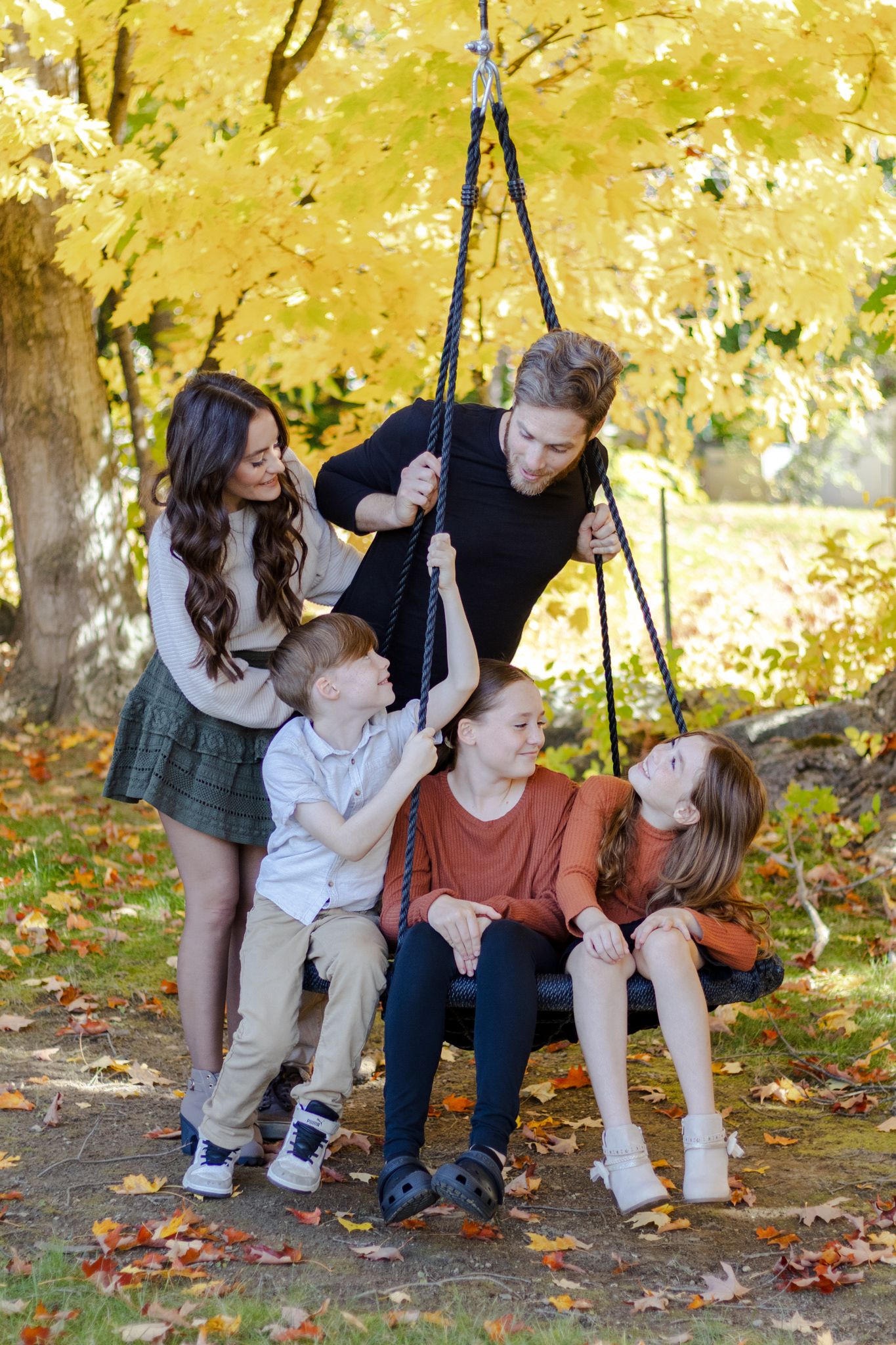 Family sits together on wooden swing surrounded by golden autumn leaves in outdoor setting.
